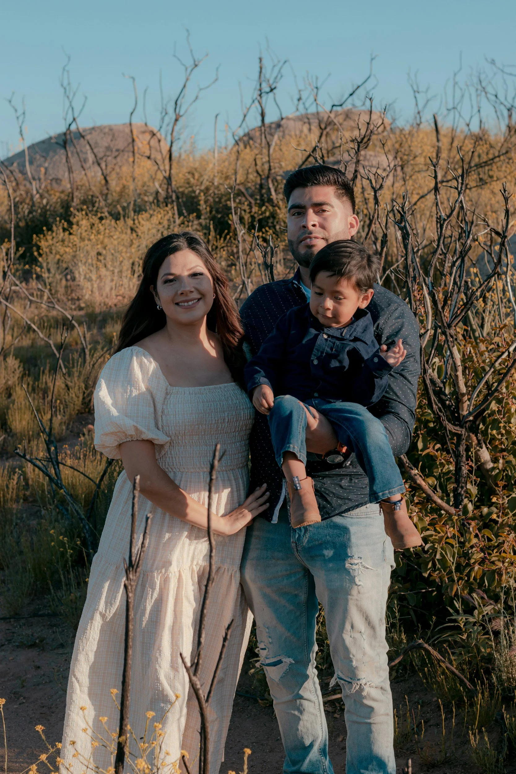 A family of three posing outdoors in a field with yellow flowering plants and leafless bushes. The woman is wearing a cream-colored dress, the man is wearing a dark blue shirt with white polka dots and light jeans, and the young boy is wearing a dark blue shirt and jeans. The background features a hilly landscape and a clear blue sky.