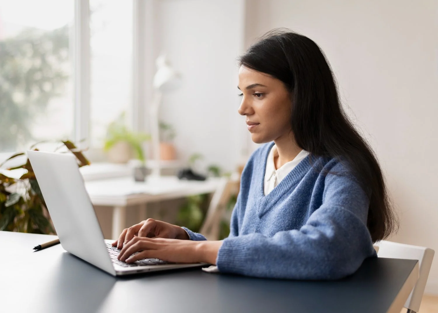 A young woman with long dark hair, wearing a blue sweater and white collared shirt, sitting at a desk working on a laptop in a well-lit room with large windows and houseplants.
