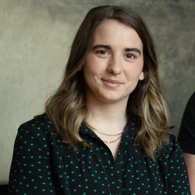 Portrait of a woman with brown hair wearing a black shirt with green patterns, smiling slightly against a neutral background.