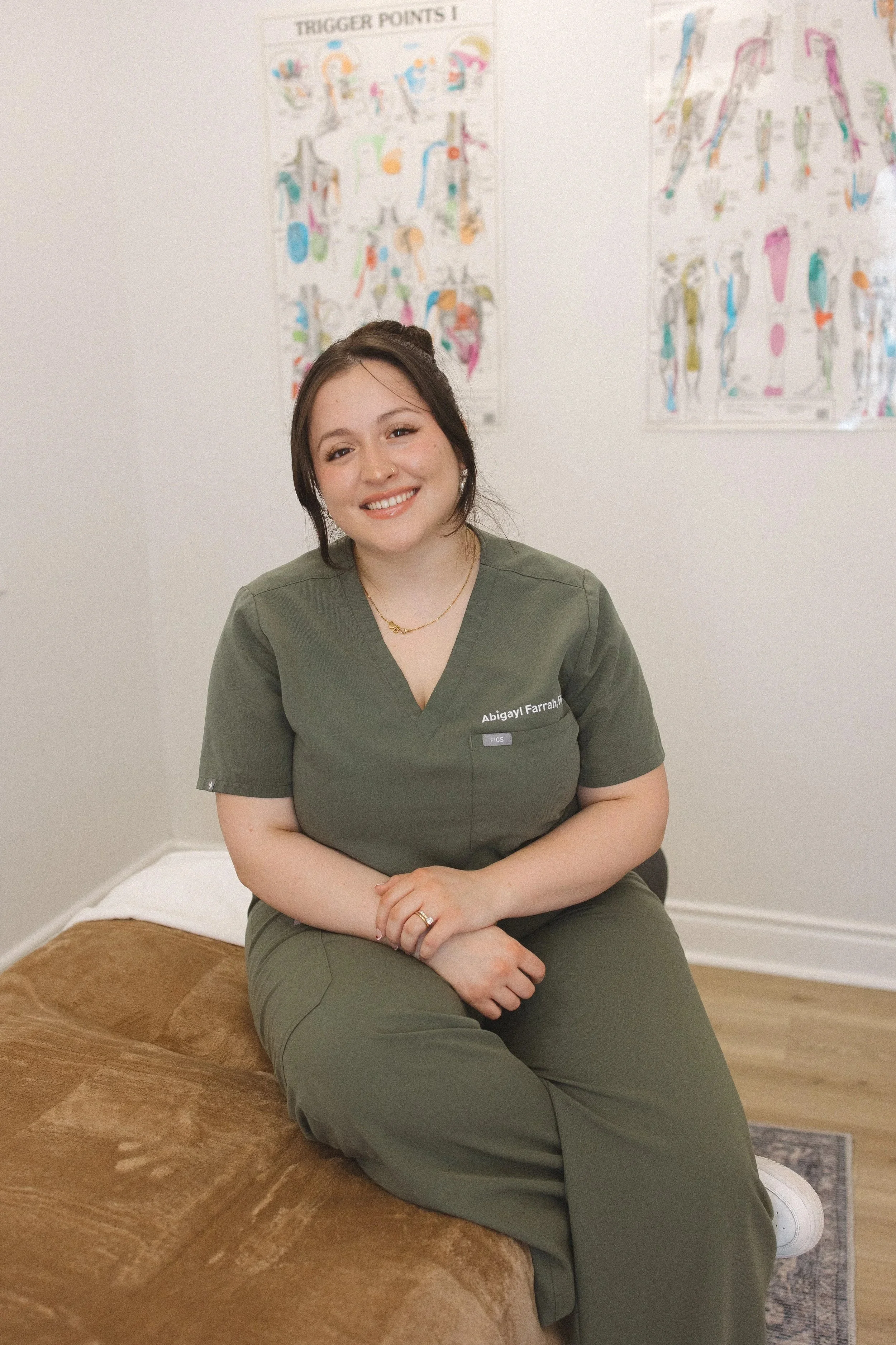 A young woman with long dark hair smiling outdoors. She is wearing black scrubs with a name tag, gold hoop earrings, and a white watch, standing near a tree and autumn leaves.
