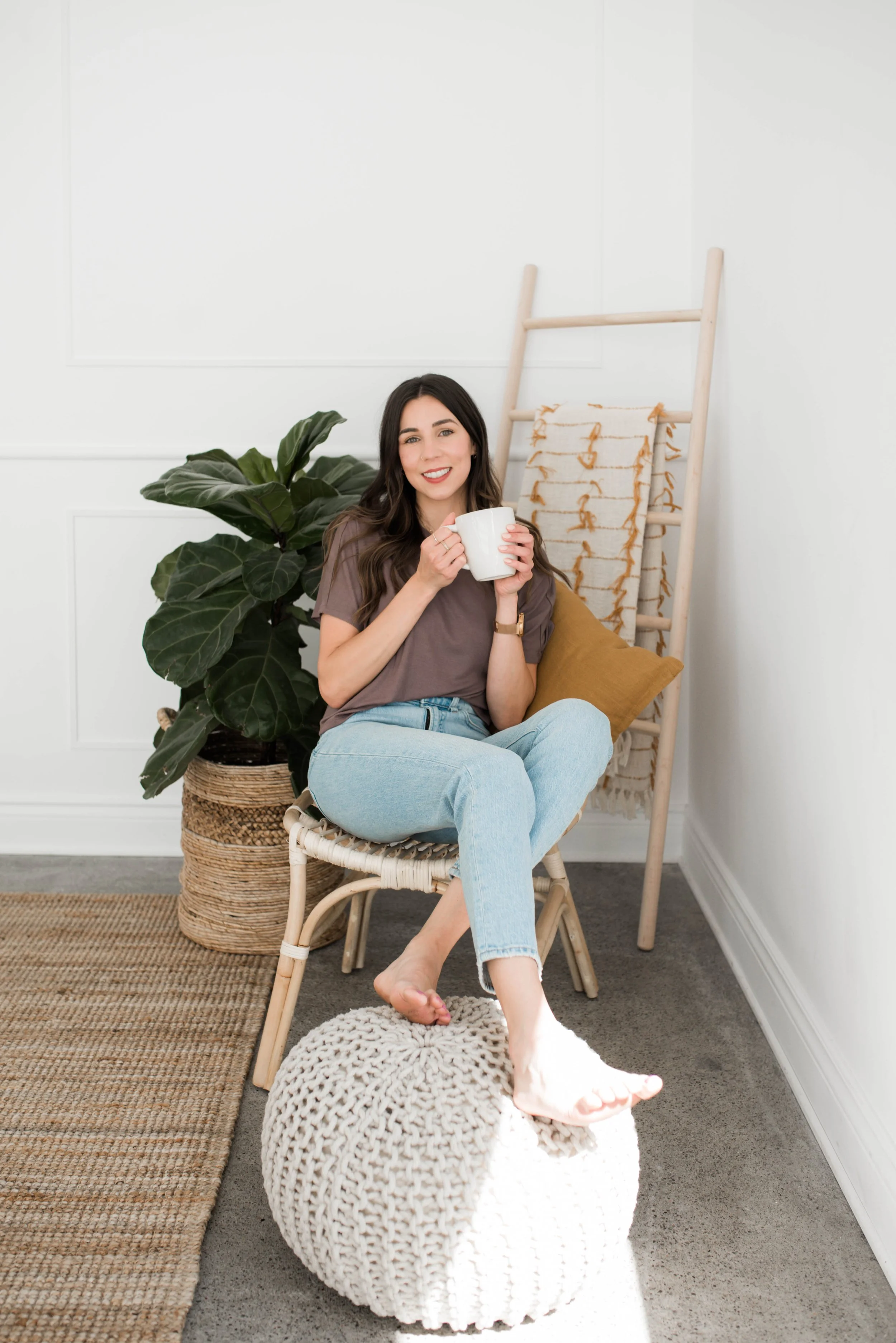 A woman sitting on a round, knitted ottoman, holding a white mug, smiling, next to a large potted plant and a wooden ladder with a blanket draped over it in a bright, minimalistic room.