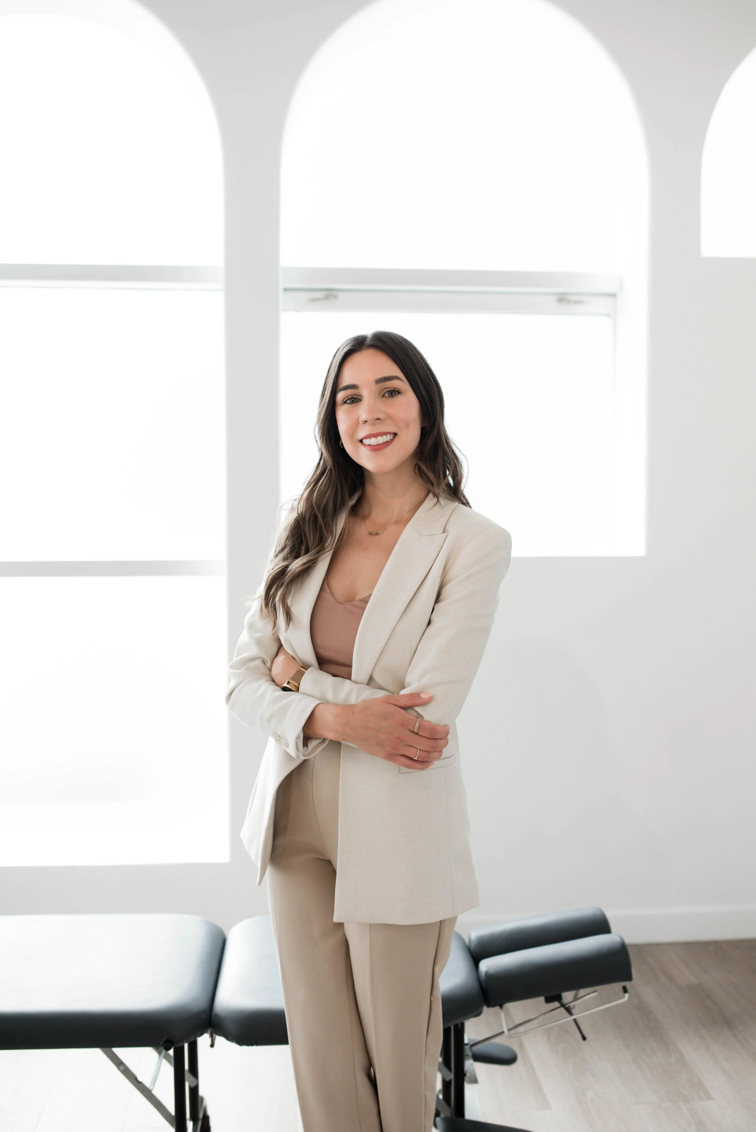 A smiling woman with long dark hair standing in a bright room with white walls and large windows, wearing a beige suit and light brown top, with her arms crossed.