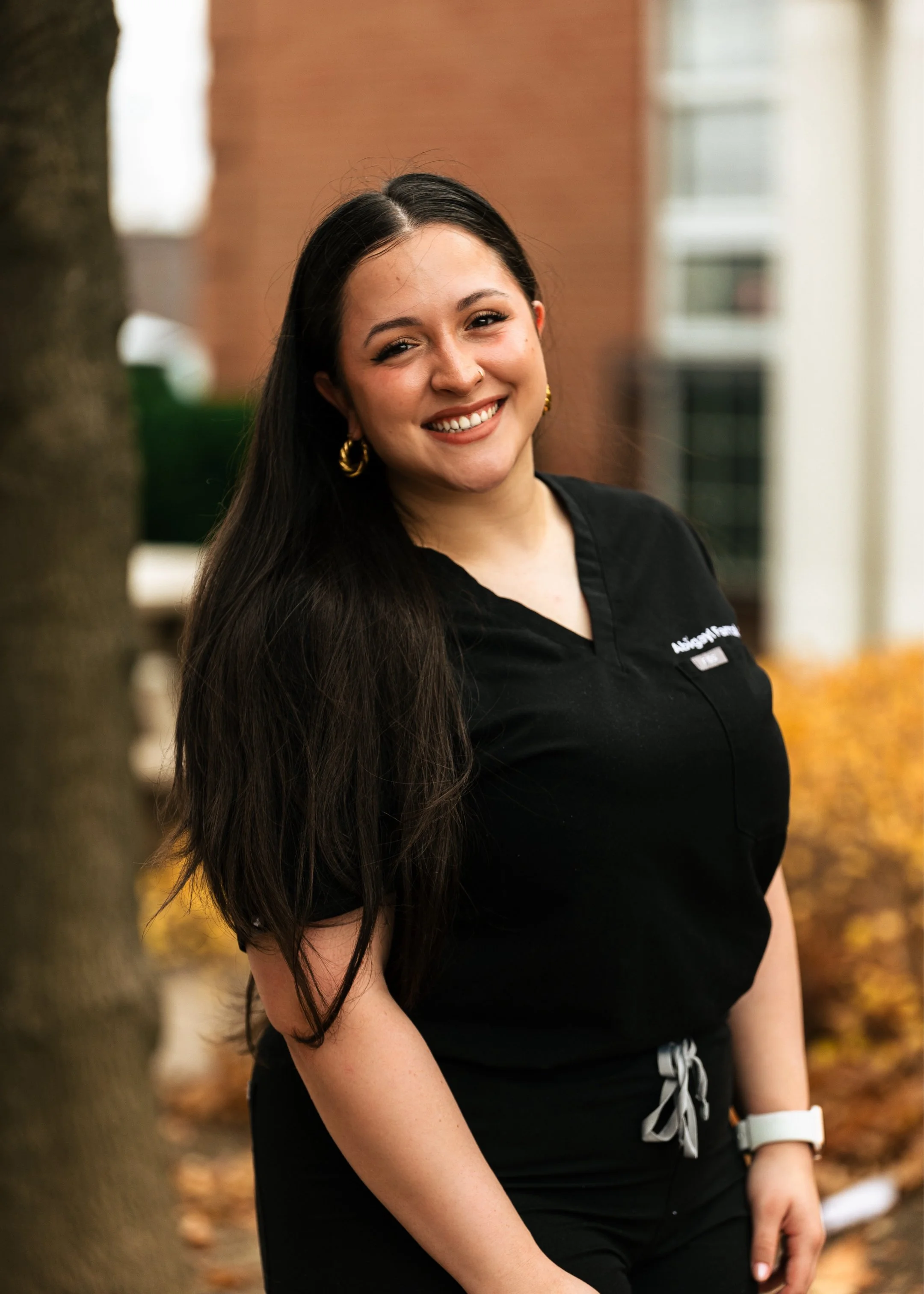 A smiling woman with long dark hair wearing black scrubs and gold hoop earrings, standing outdoors in front of trees and a brick building.