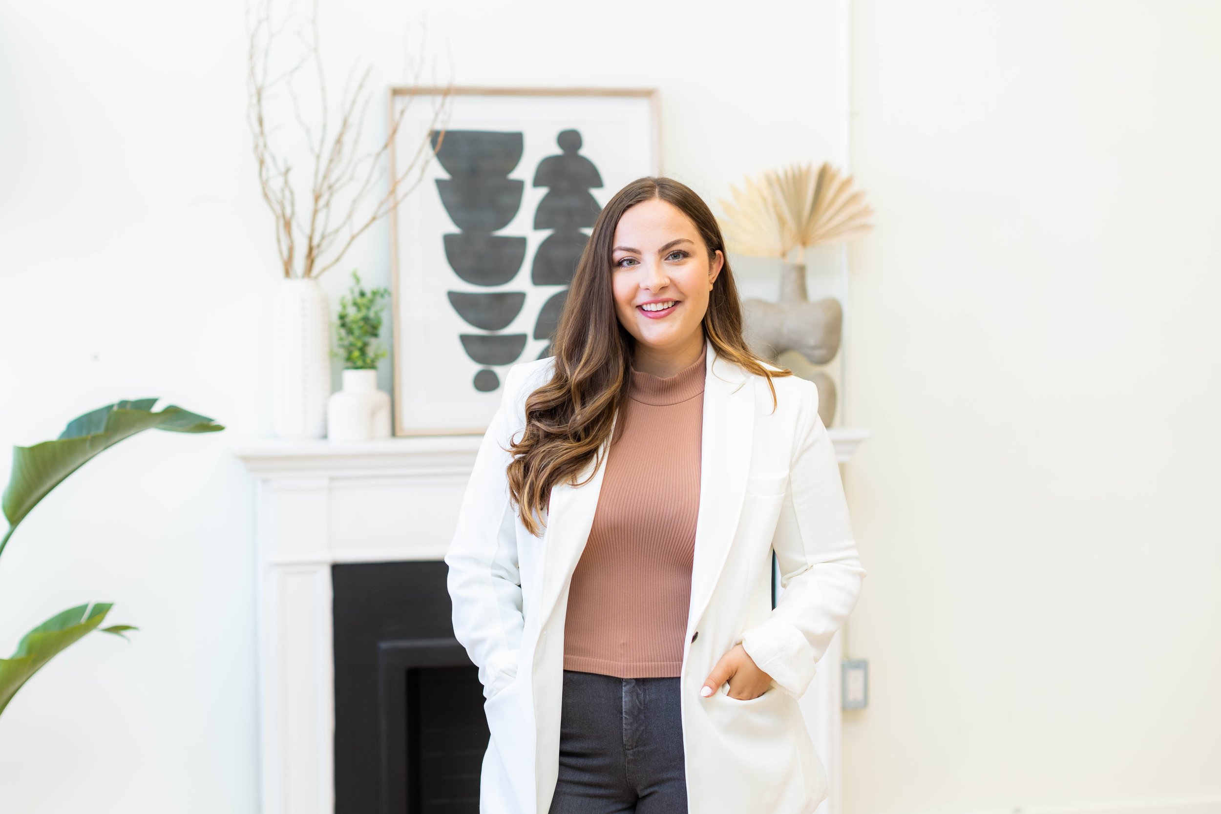 Woman with long hair and glasses wearing a white blazer and brown top leaning against a wall, smiling.
