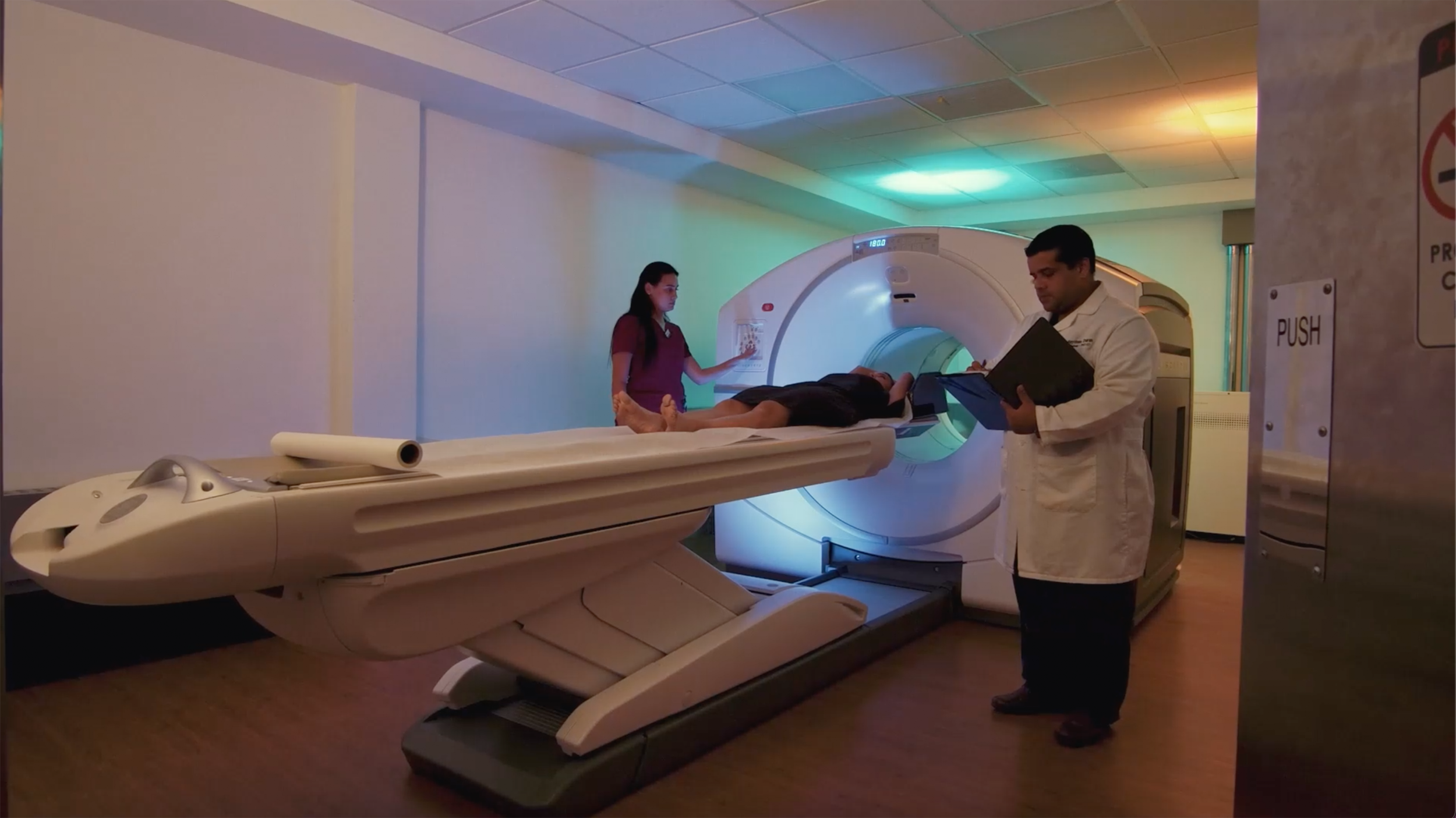 Paciente acodo en una máquina de resonancia magnética, con un técnico y otra persona observando en la sala de escaneo.