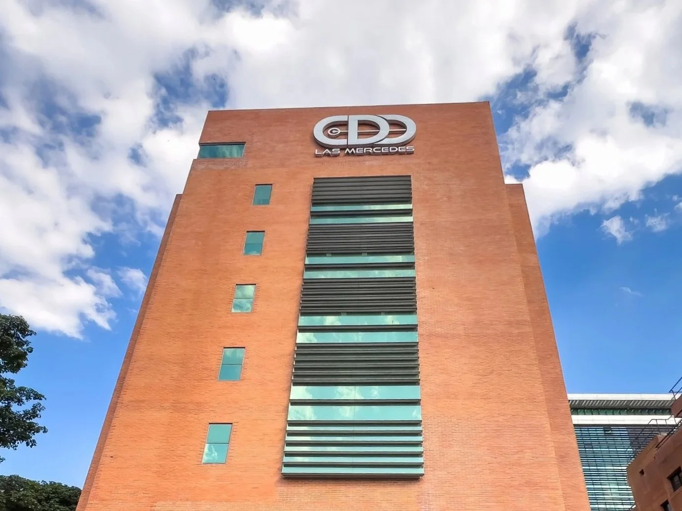 Edificio alto de ladrillos con ventanales de vidrio y una señal que dice 'Las Mercedes' en la parte superior, con cielo azul y nubes al fondo.