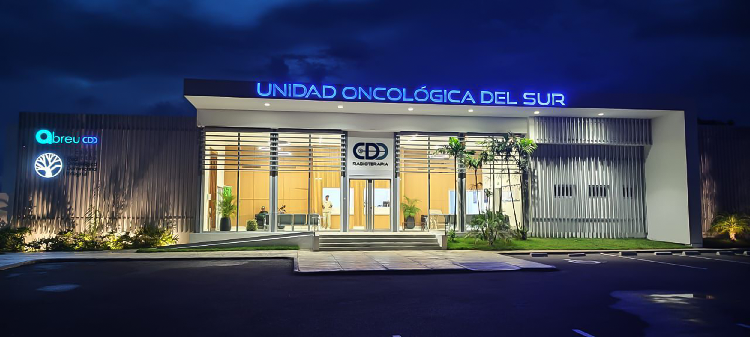 Edificio de un centro oncológico y radioterapia iluminado de noche, con el cartel que dice 'Unidad Oncológica del Sur' y plantas en el exterior.