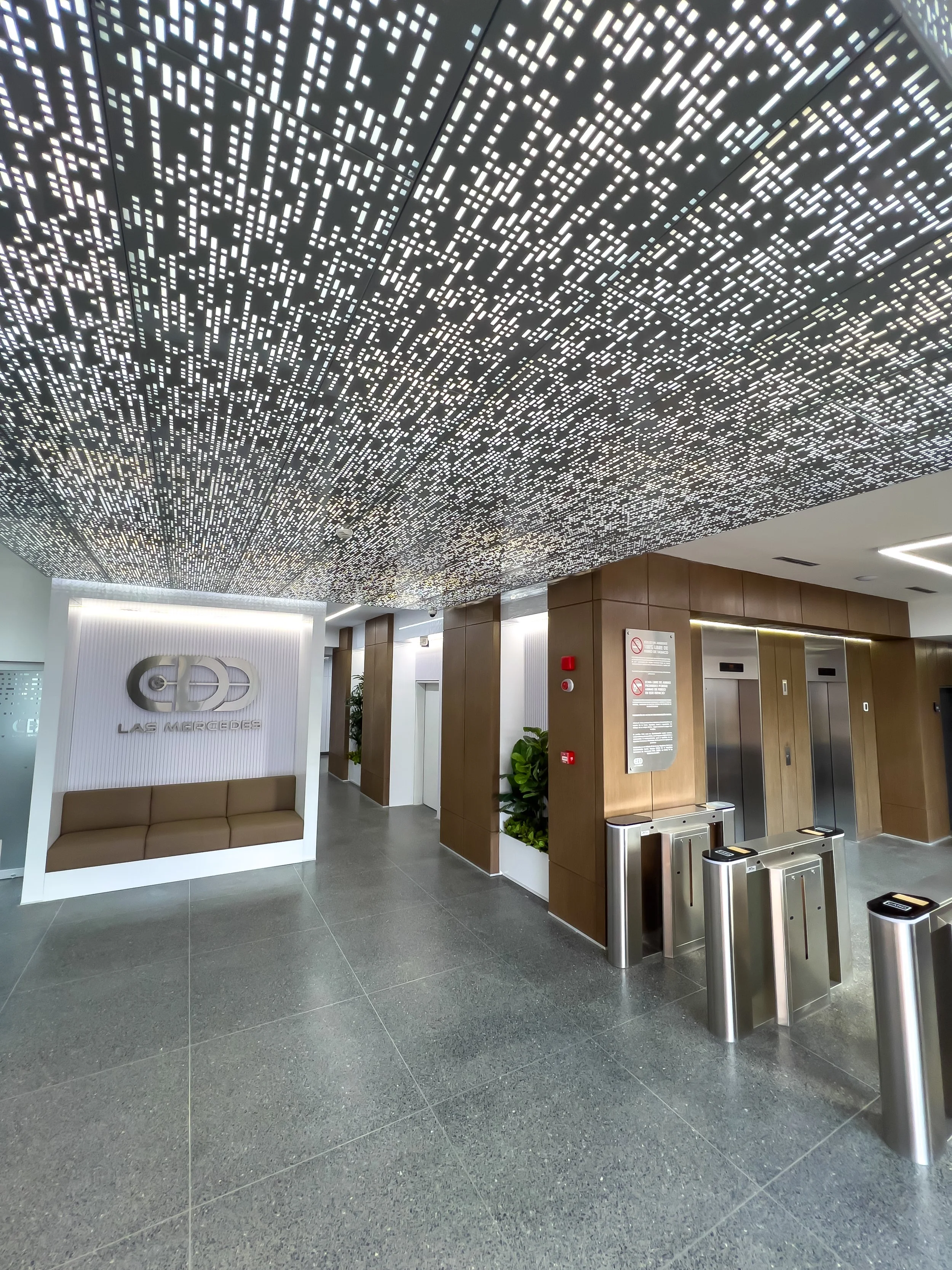 Vista del vestíbulo de un edificio con suelo y paredes de color gris oscuro, con puertas de ascensor y torniquetes de control de acceso. En el techo, un diseño artístico de luces LED en patrón de líneas y cuadros blancos. También hay un letrero en la