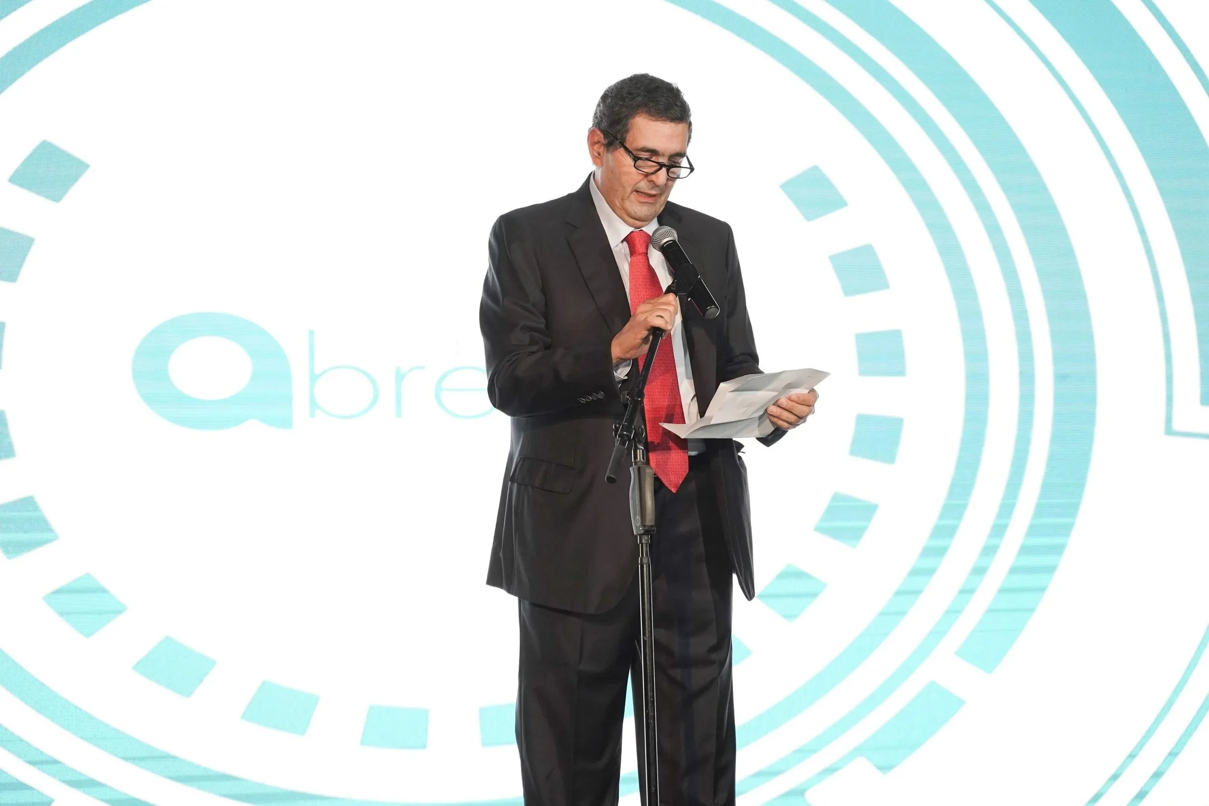 Hombre con traje negro, corbata roja y lentes leyendo un papel, frente a un micrófono en un escenario con fondo azul y diseños geométricos.