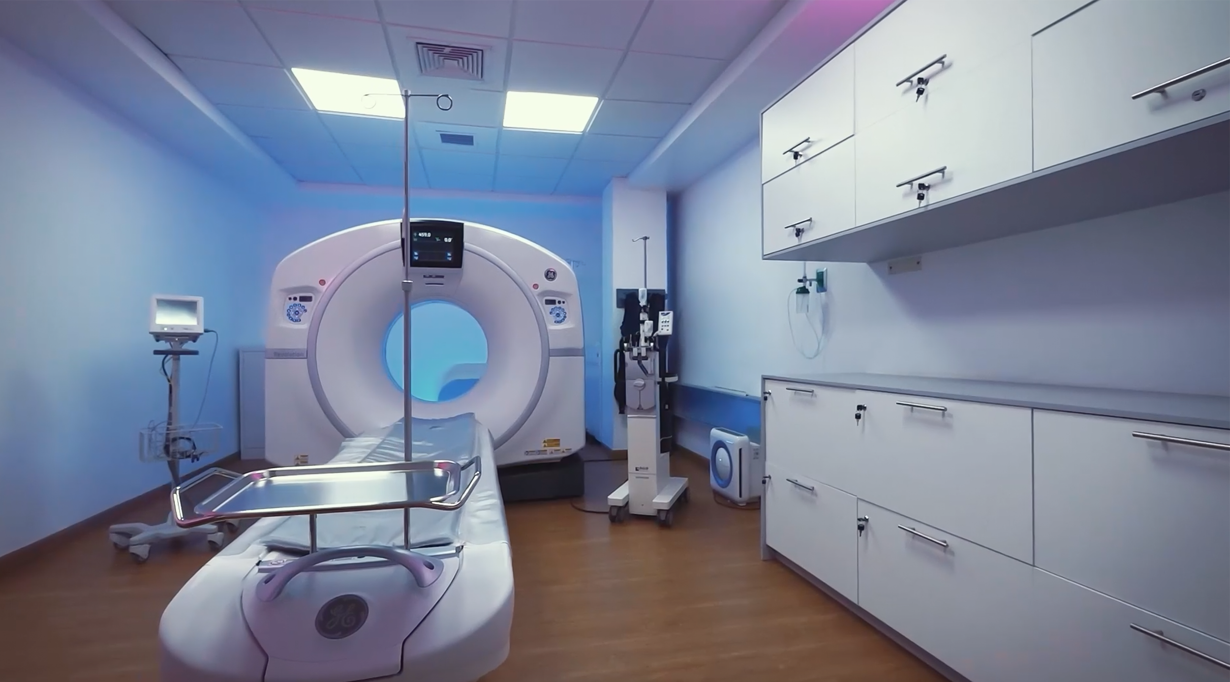 Sala de radiología con máquina de tomografía computarizada, cama para paciente, monitores, gabinetes y equipo médico en una clínica o hospital.