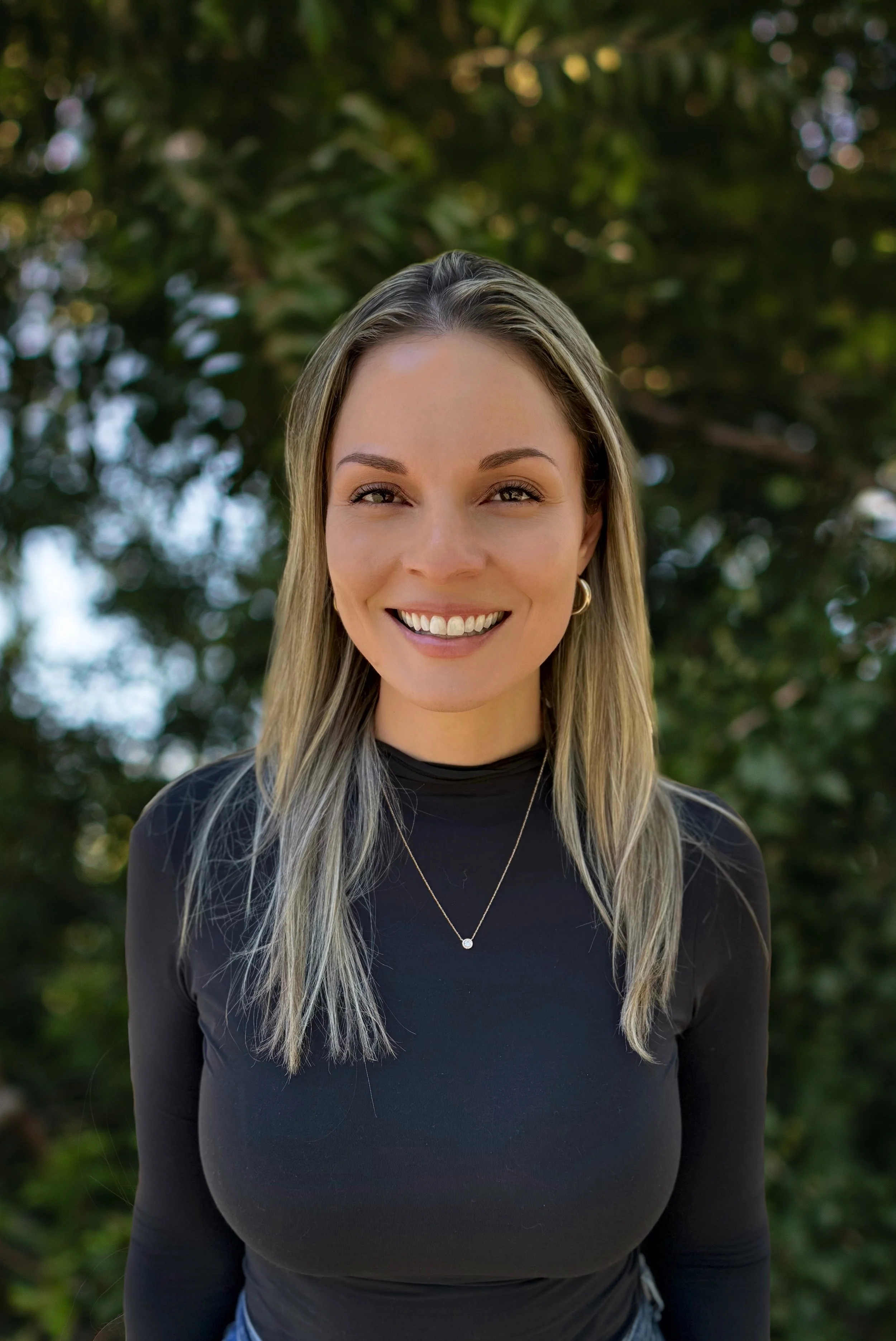 A smiling woman with wavy blonde hair wearing a black turtleneck and jewelry, standing outdoors in front of green trees.