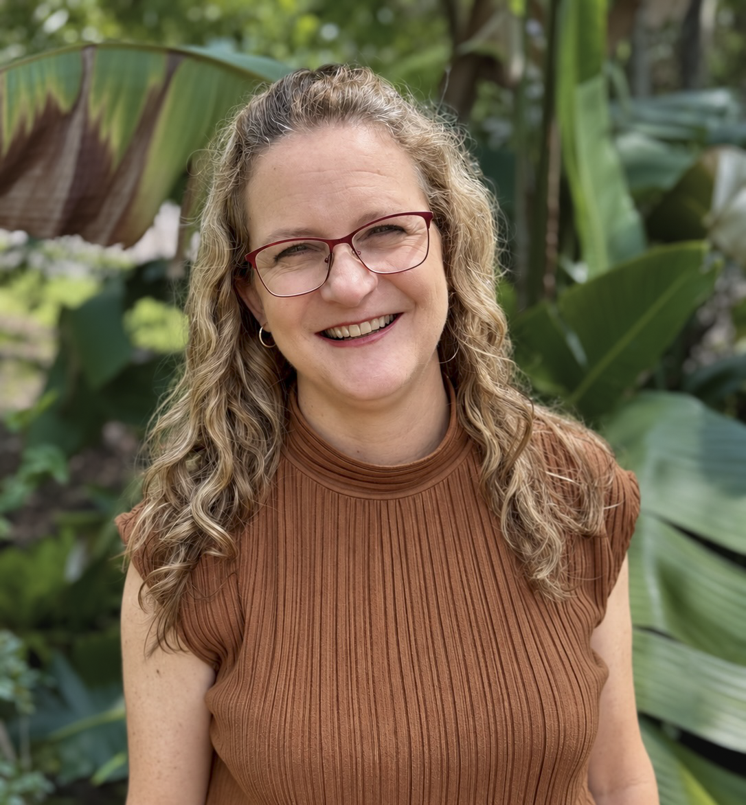 A woman with wavy, shoulder-length hair and glasses, smiling outdoors with greenery in the background, wearing a denim jacket over a rust-colored top.