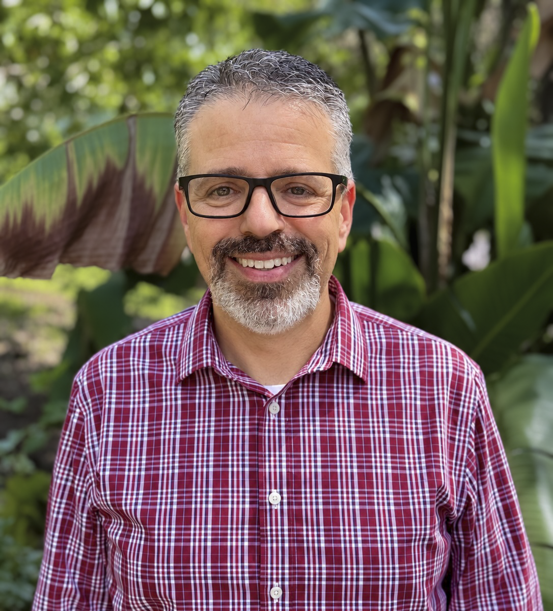 A middle-aged man with glasses, gray hair, and a beard, smiling outdoors with a background of green trees.