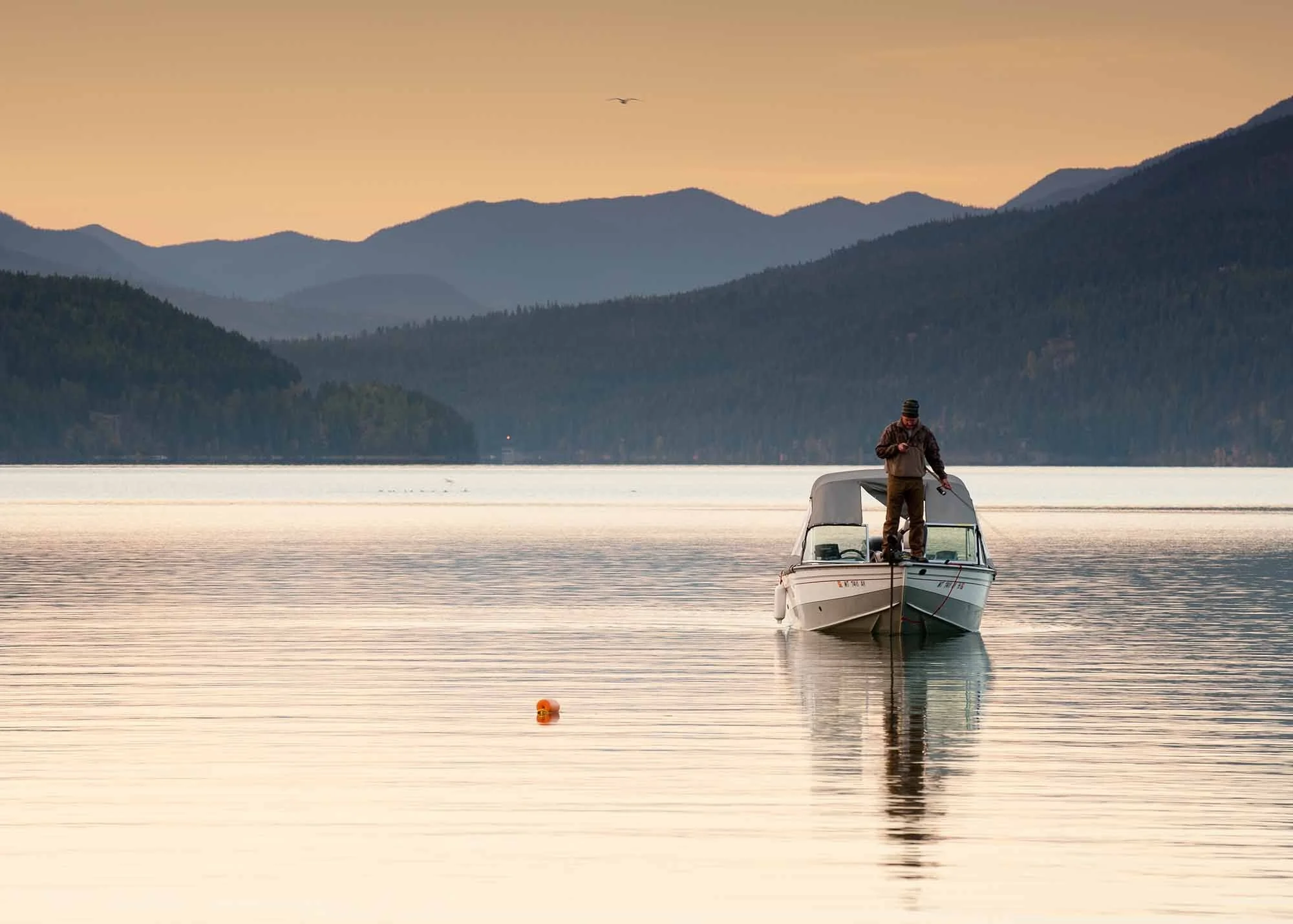 Man standing on the prow of a boat on Whitefish Lake, with mountains in the background at sunset