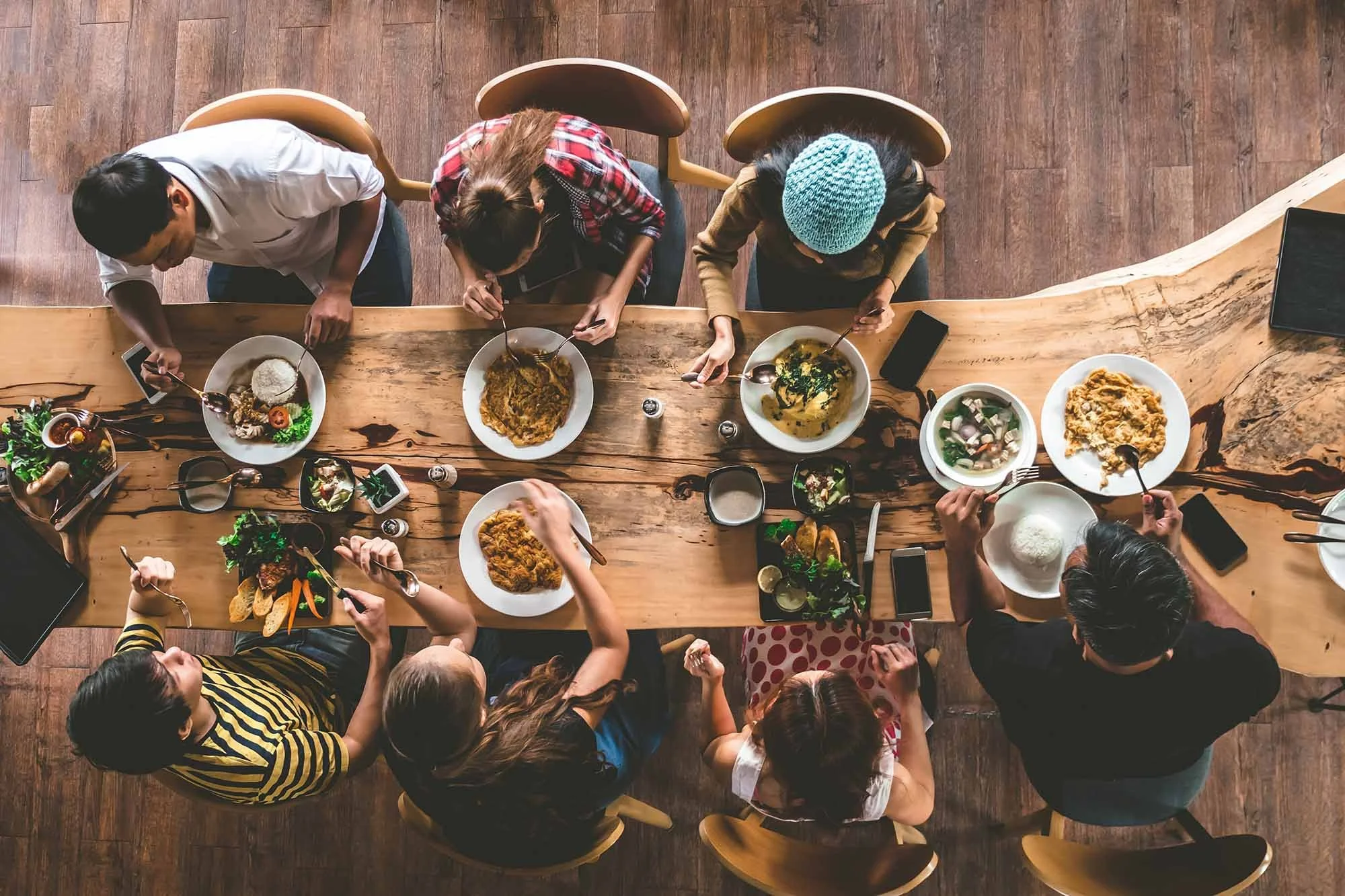 Aerial view of friends sitting at a reclaimed wood dining table, with multiple plates of food on the table