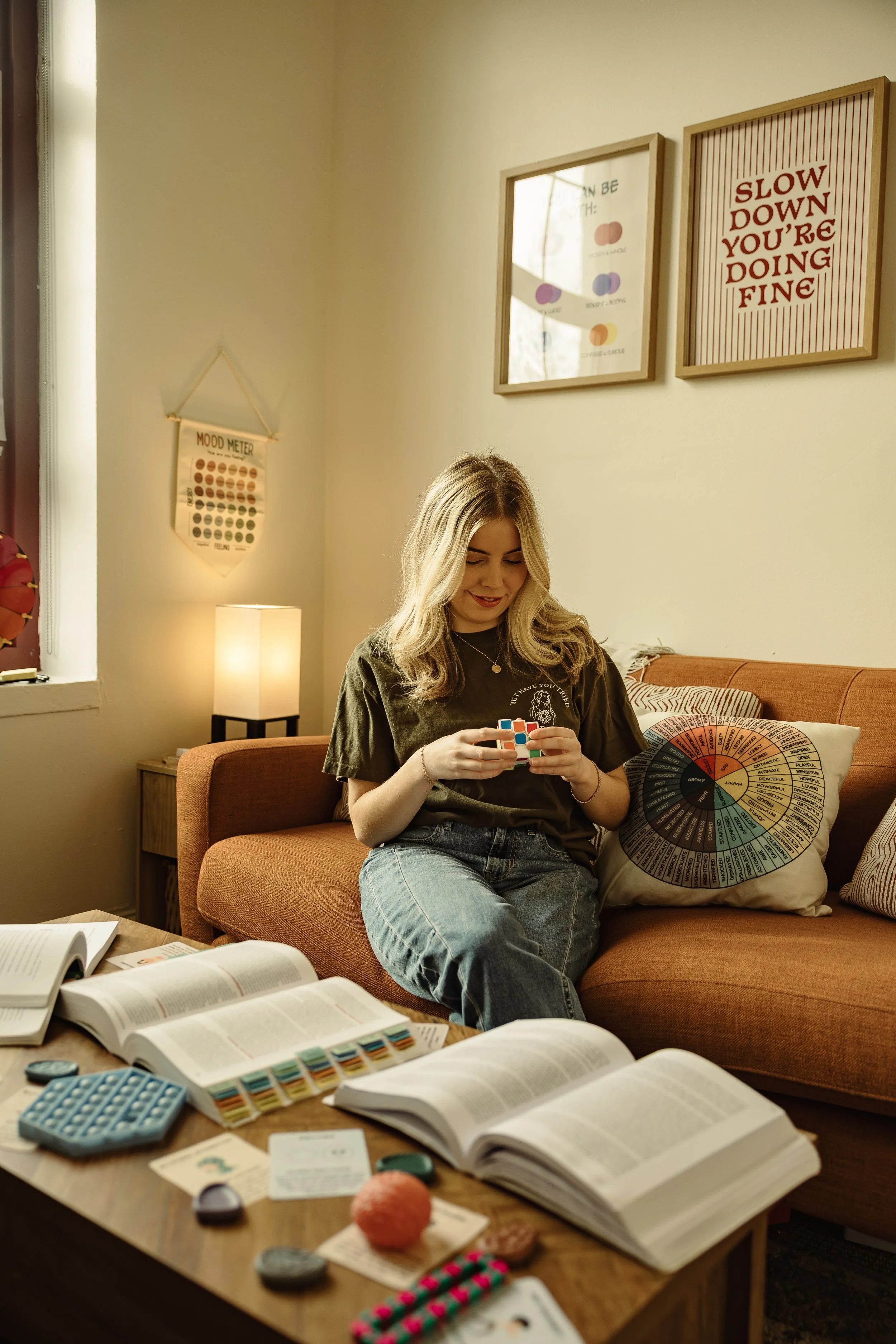A young woman with long blonde hair sitting on a couch, holding a Rubik's cube in her hands, surrounded by open books, notebooks, and various craft supplies on a coffee table. The room has a warm, cozy atmosphere with a lamp, framed posters on the wall, and a decorative pillow with a colorful wheel of emotions.