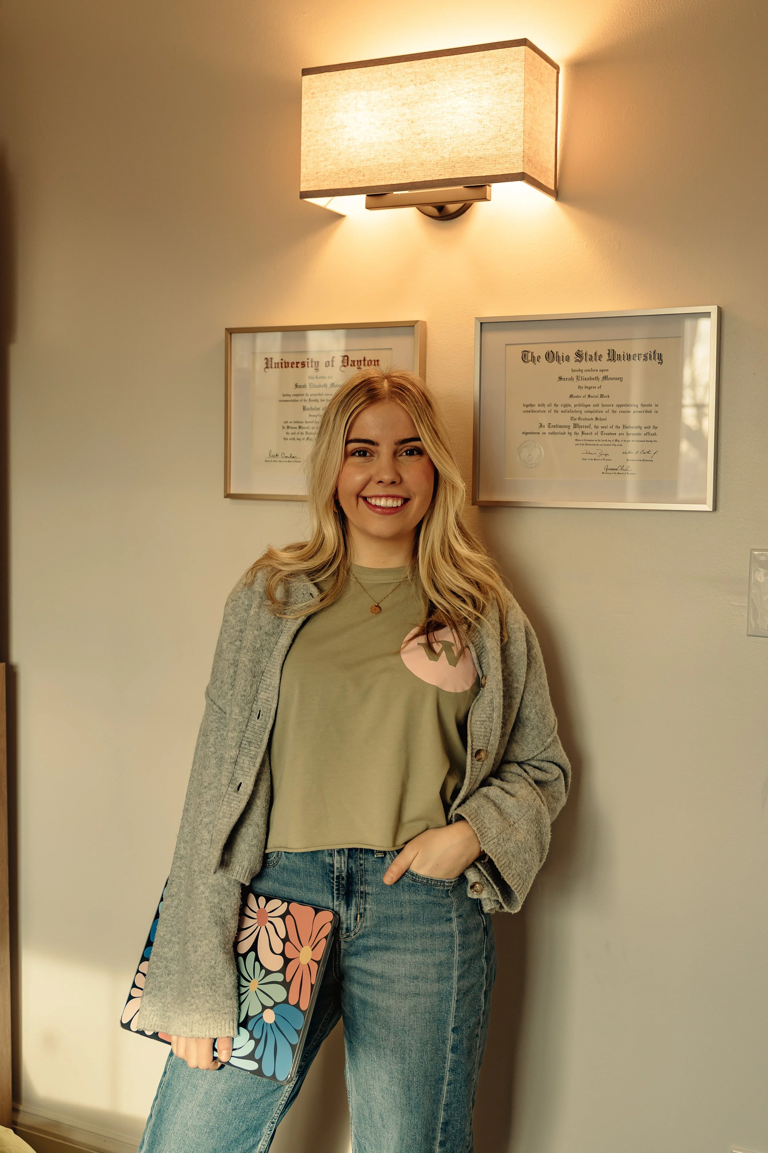 A young woman with blonde hair and a friendly smile, dressed in a green shirt, gray cardigan, and jeans, standing indoors against a beige wall with framed diplomas and a wall-mounted light fixture.