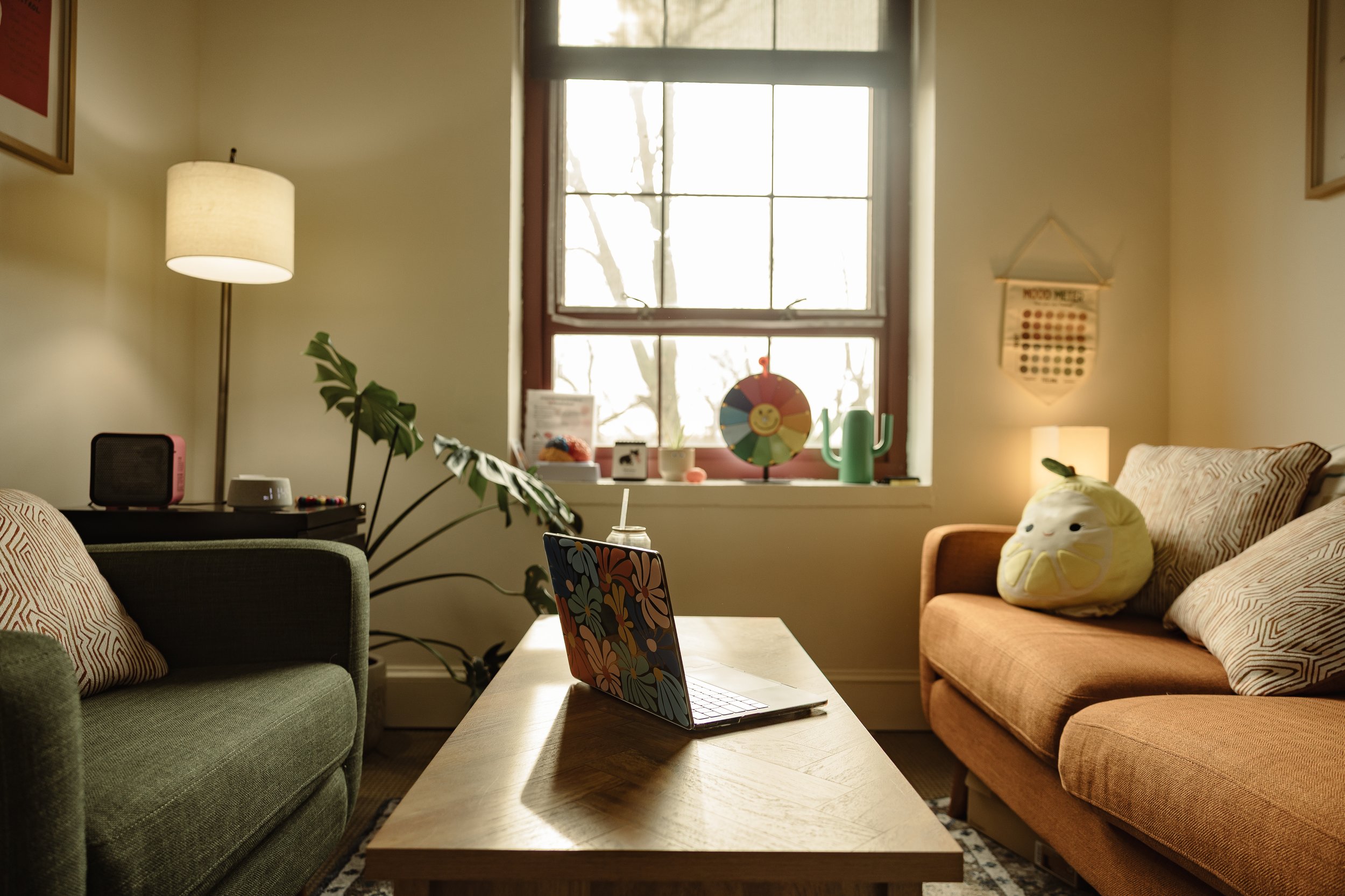 A cozy living room with a green sofa on the left and a peach-colored sofa on the right, both with patterned pillows. A wooden coffee table in the center holds a colorful laptop. Behind the coffee table, a large window with tree branches outside lets in natural light. The windowsill has various decorative items, including a green watering can and a circular rainbow-colored decoration with a smiley face. To the left, a tall lampshade and a potted plant are visible, while on the right wall, there is a small wall hanging and a lit table lamp.