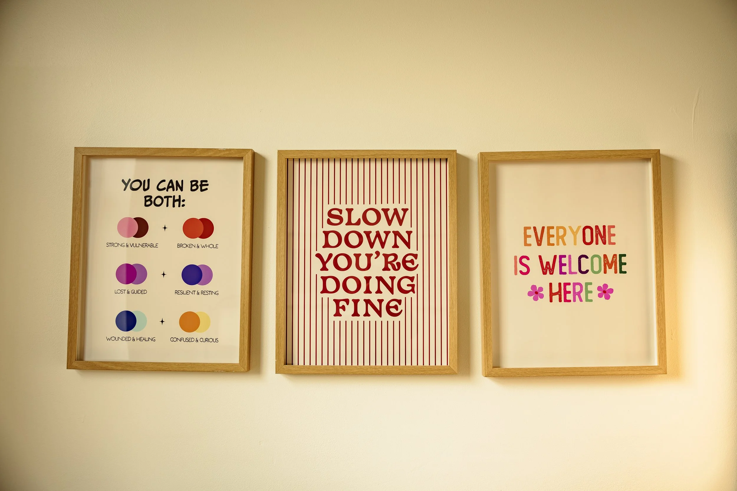Three framed posters on a wall with inspirational messages and colorful graphics. The left poster shows color-coded pairs with descriptive words, the middle poster reads 'Slow down you're doing fine' in red text on a striped background, and the right poster has multicolored text saying 'Everyone is welcome here' with small flower graphics.