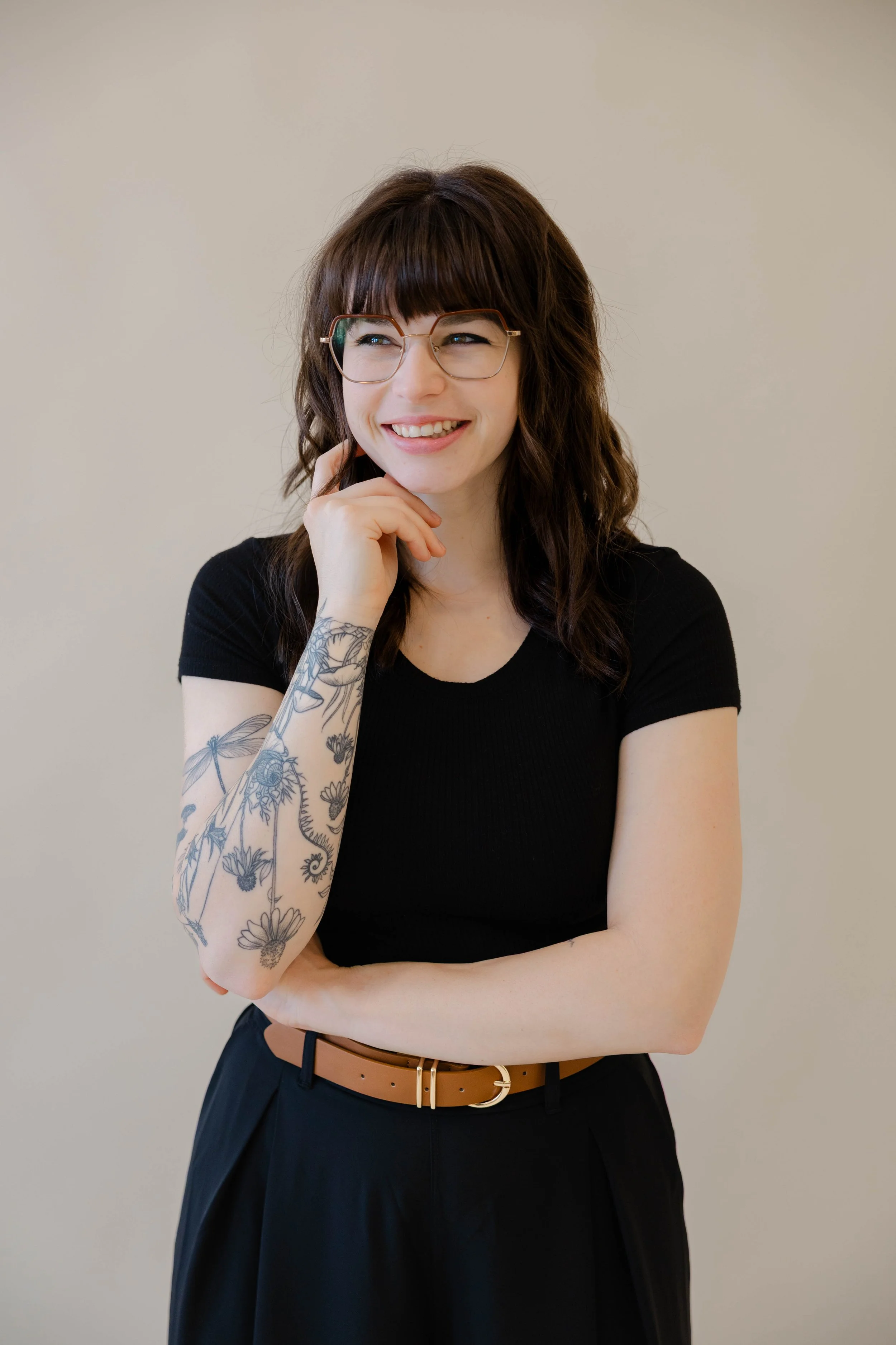 A woman with shoulder-length brown hair, glasses, and tattoos on her left arm, wearing a black top and a tan belt, smiling thoughtfully with her right hand on her chin and crossed arms against a plain beige background.