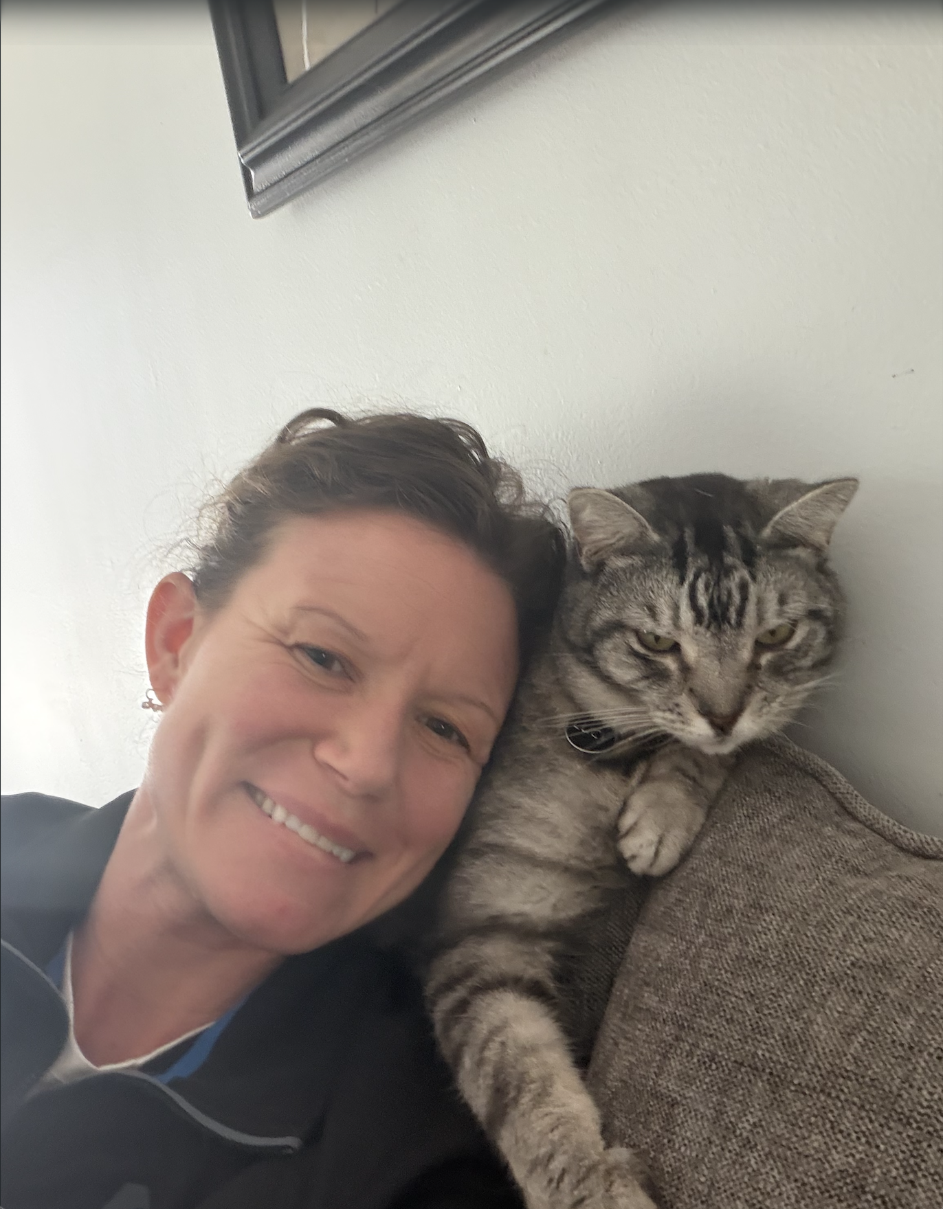 A woman and a tabby cat lying close together on a couch, smiling and looking at the camera.