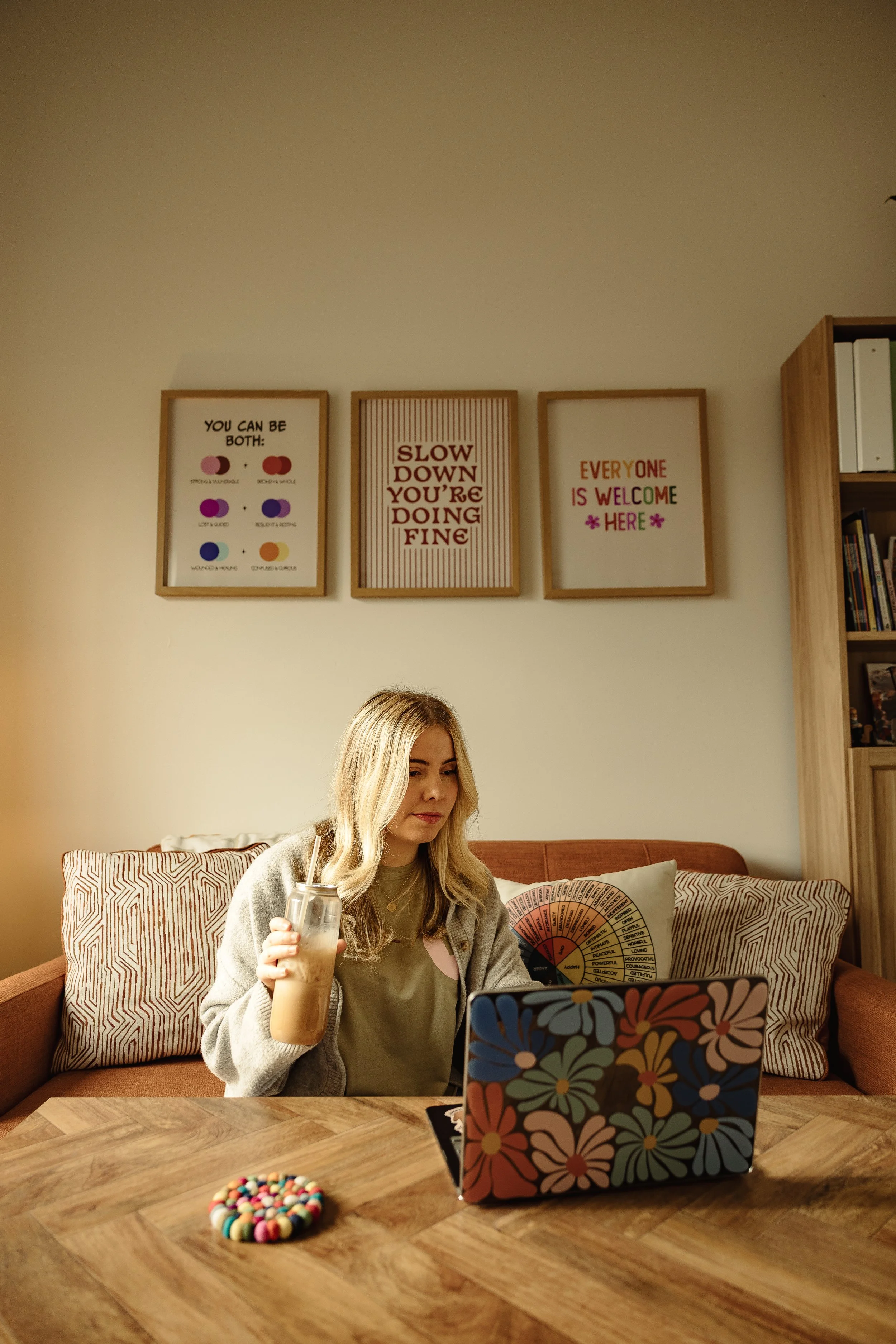 A woman with long blonde hair sitting on a brown couch with patterned pillows, holding a glass of iced coffee, in a cozy room with wooden furniture and framed quotes on the wall. She is using a colorful floral-patterned laptop on a wooden table, with a small colorful round object on the table.
