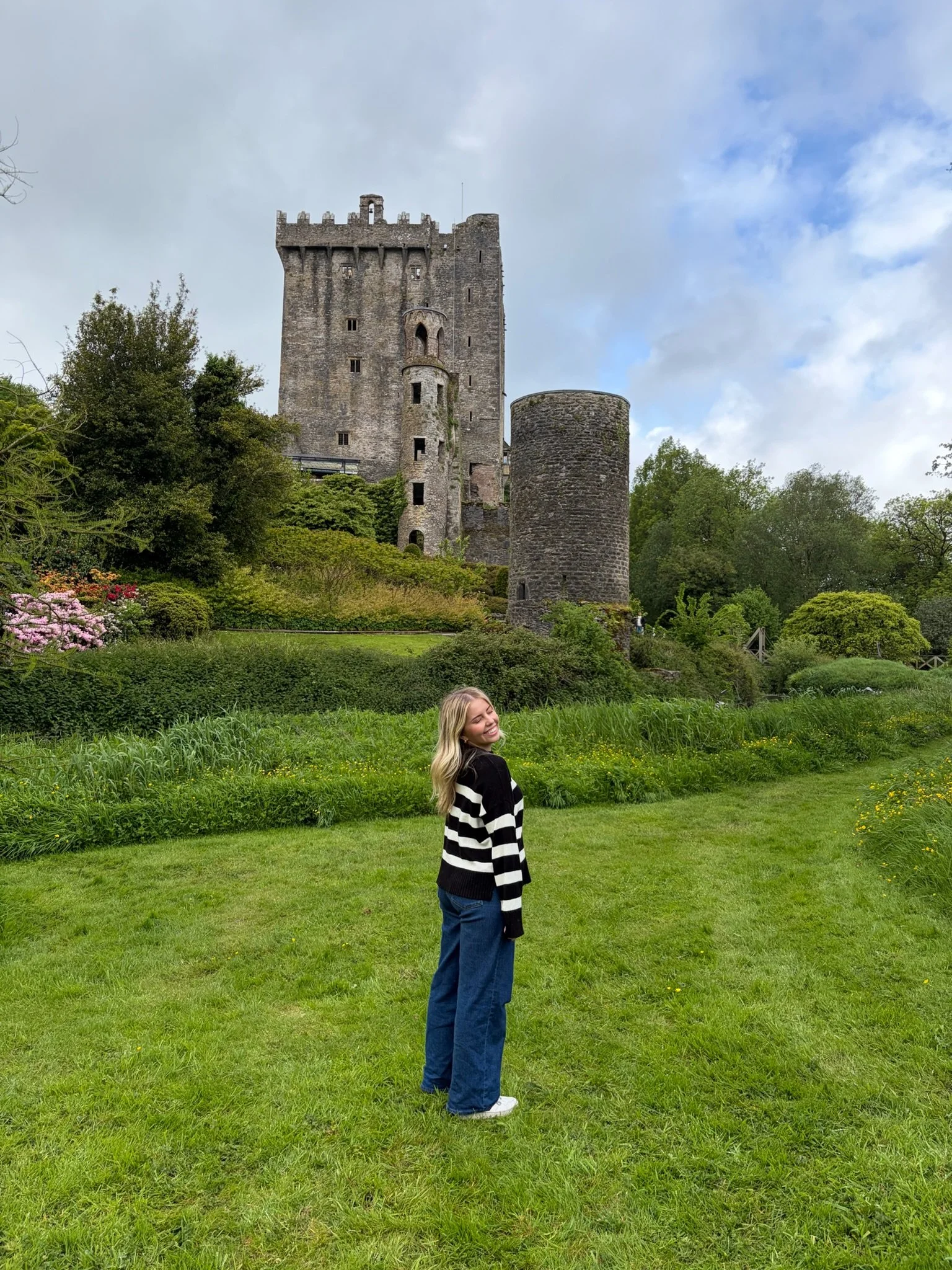 A young woman with blonde hair, wearing a black and white striped sweater, blue jeans, and white sneakers, standing on a grassy lawn with a large stone castle and trees in the background, smiling with her eyes closed.