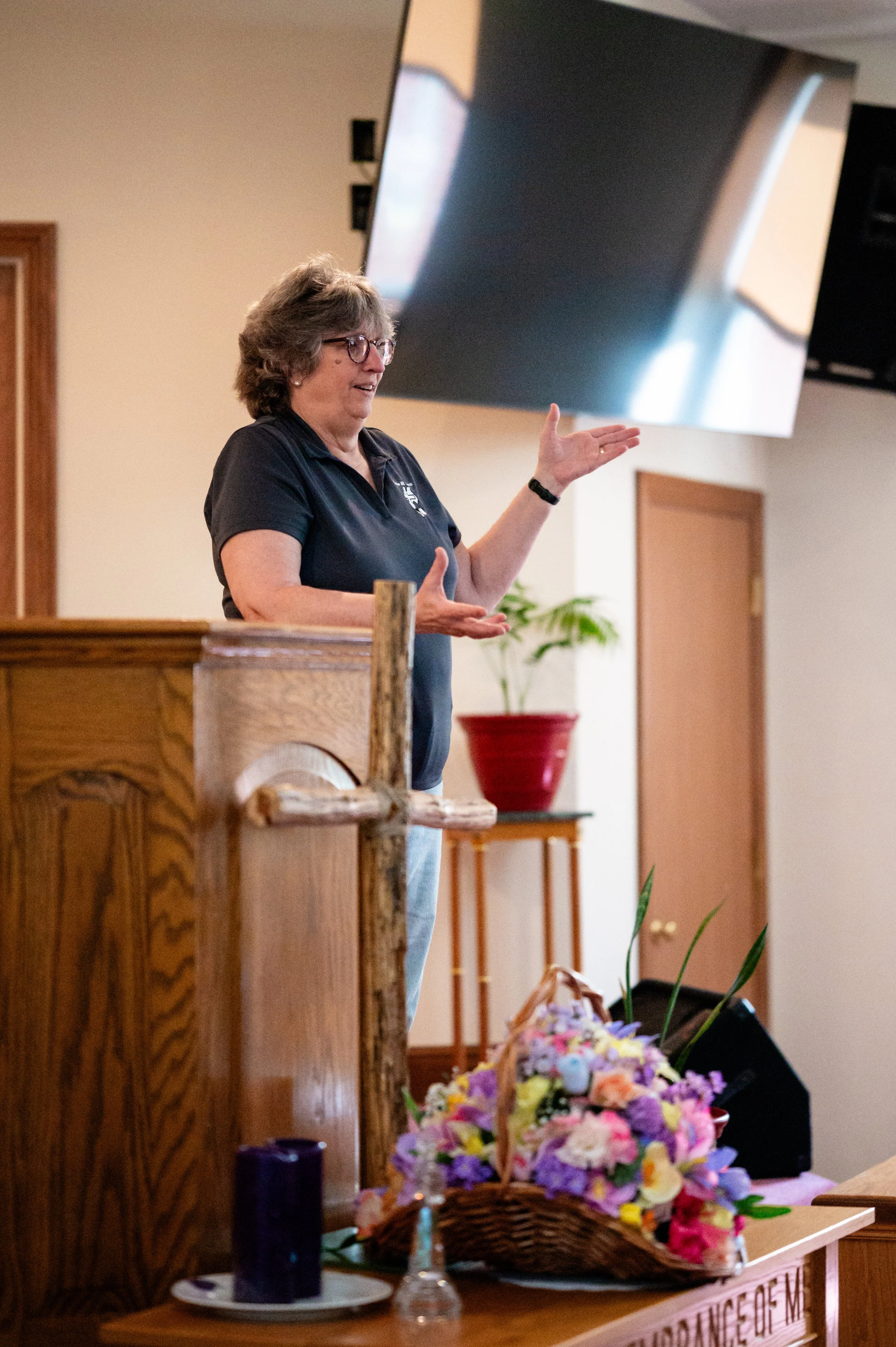 A woman wearing glasses and a black polo shirt stands behind a wooden podium, gesturing with her right hand. In front of her on the table, there is a flower arrangement in a basket, a purple candle, a small glass bottle, and a white plate. The background includes a flat-screen TV mounted on the wall and a red flower pot with a green plant.