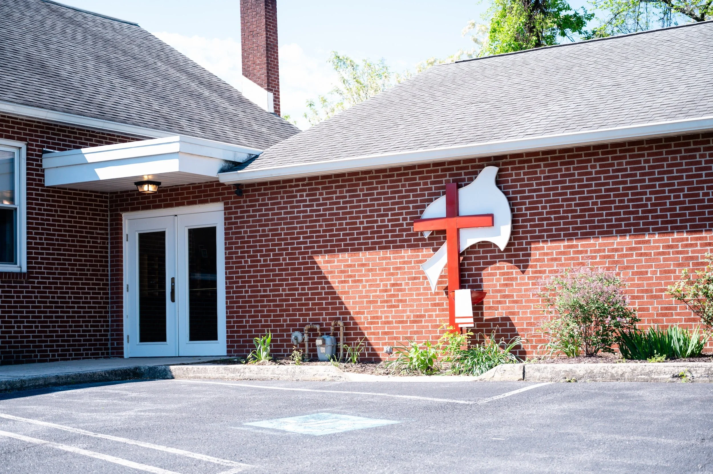Red brick building with a white door, windows, and a church cross sign outside. Parking lot in foreground with marked parking spaces and a small garden area.
