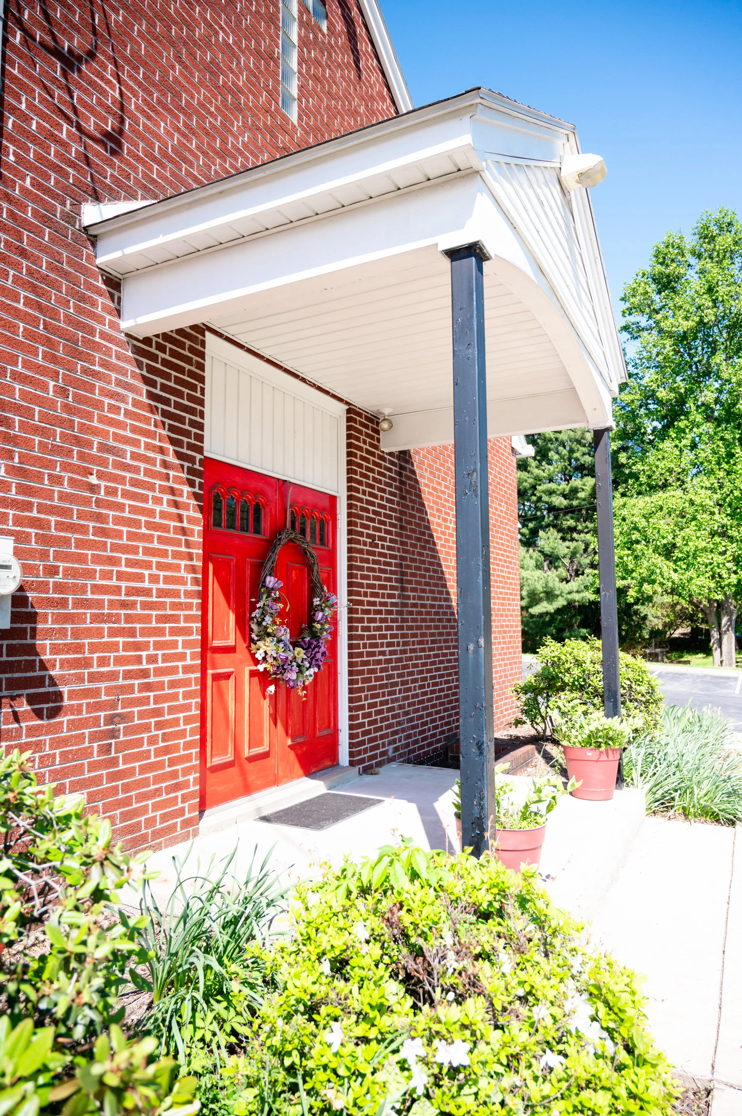 Red front door decorated with a flower wreath, set in a brick house with a white porch roof supported by black columns, surrounded by potted plants and greenery under a clear blue sky.