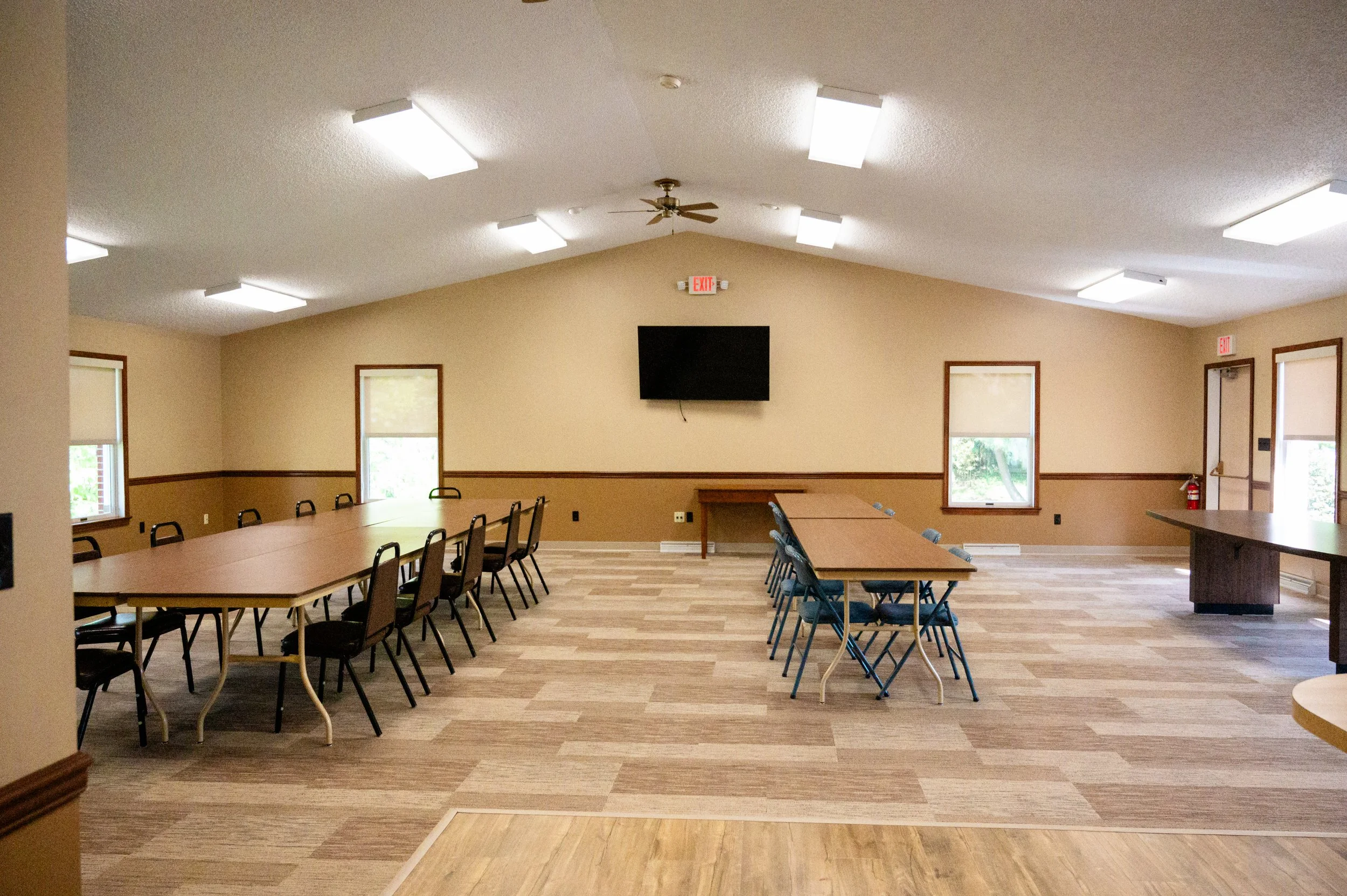 A spacious meeting or conference room with beige walls, wooden trim, and large windows. There are long tables with black and blue chairs arranged around them, and a wall-mounted TV at the front. The ceiling has white fluorescent lights and a ceiling fan. Fire extinguisher and exit signs are visible.