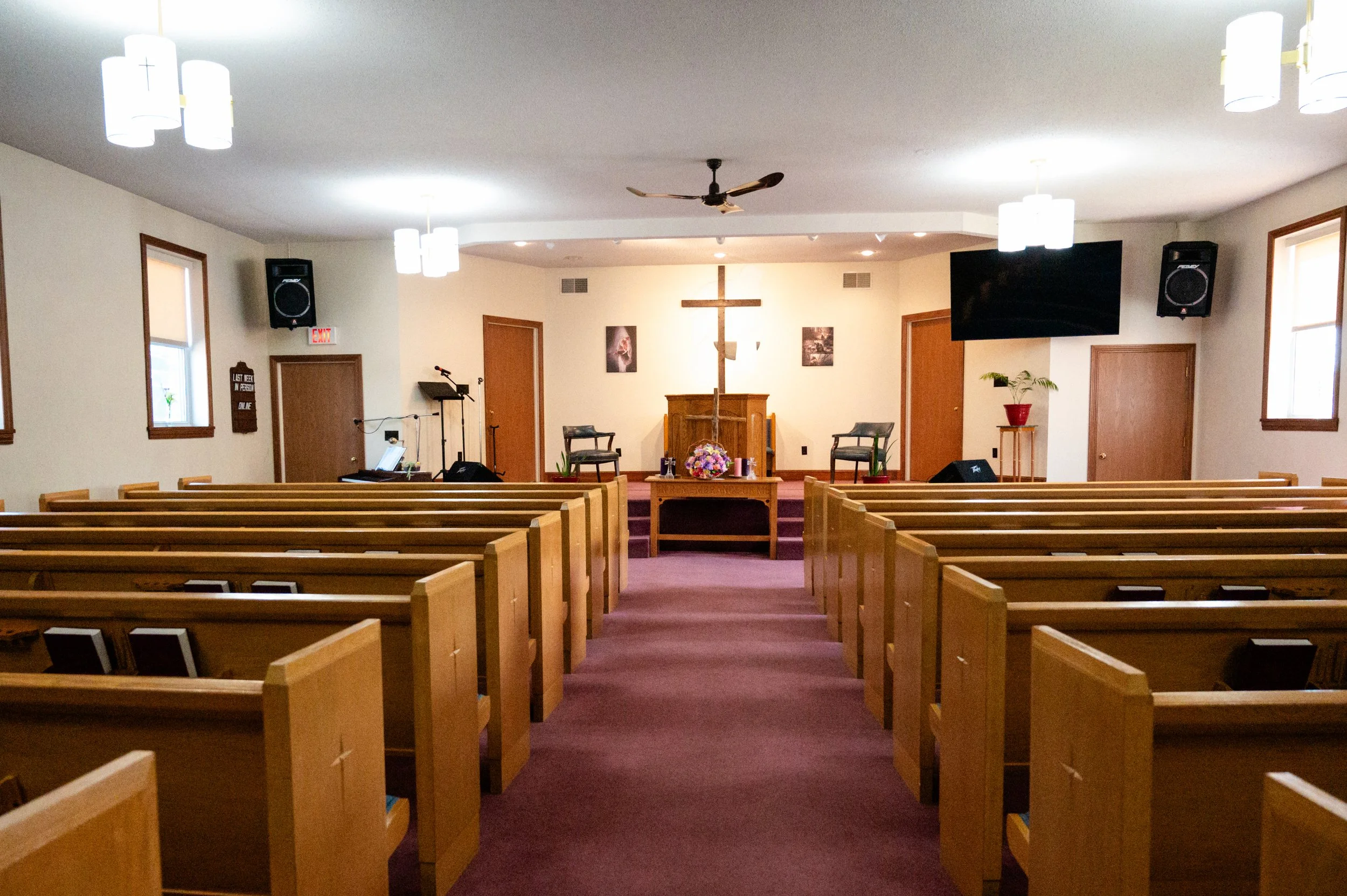 Interior view of a church sanctuary with wooden pews, a cross at the front, two chairs, a pulpit, and musical equipment, decorated with flowers and framed pictures.