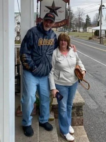 A man and woman standing outside on a sidewalk, with the man wearing an Army sweatshirt and the woman holding a purse, next to a wooden bench, in a small town setting.