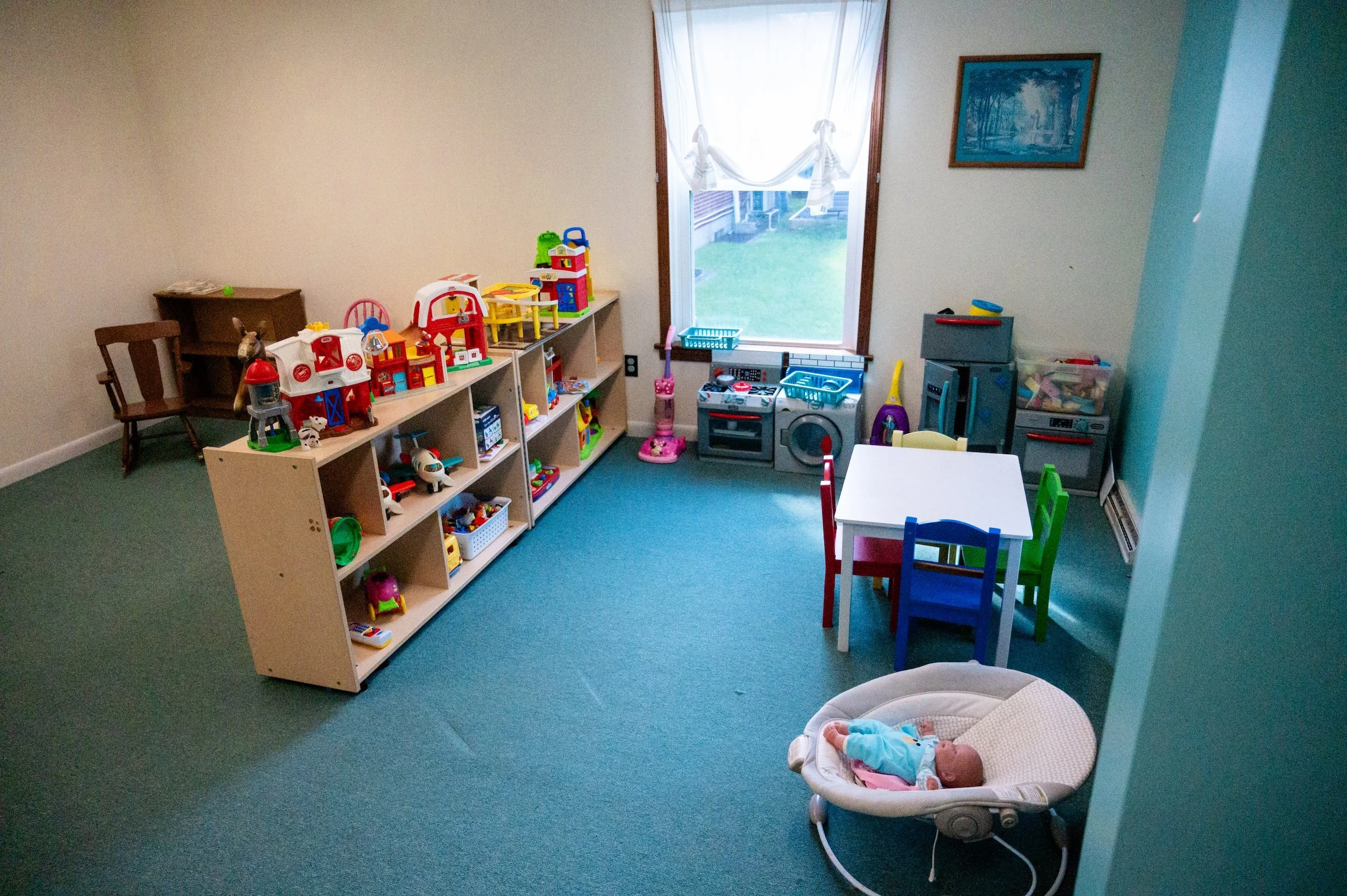 Children's playroom with toy shelves, small tables and chairs, a toy stove, and a baby doll in a bouncer, with a window showing a grassy outdoor area.