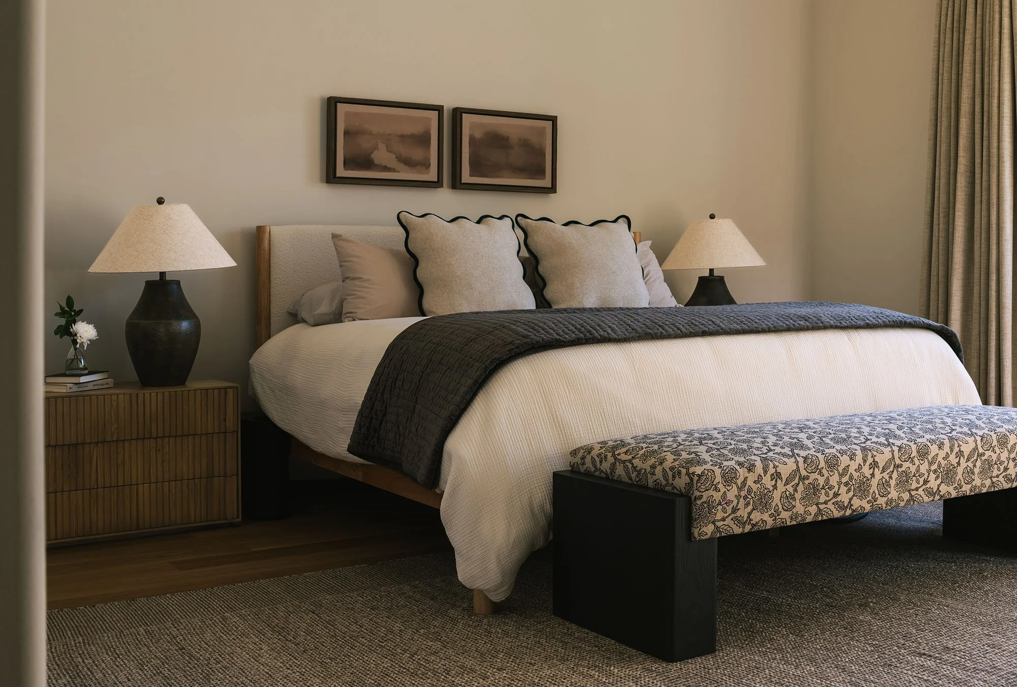 Dallas primary bedroom featuring a light upholstered bed, fluted wood nightstands, sculptural black table lamps, and a patterned upholstered bench.