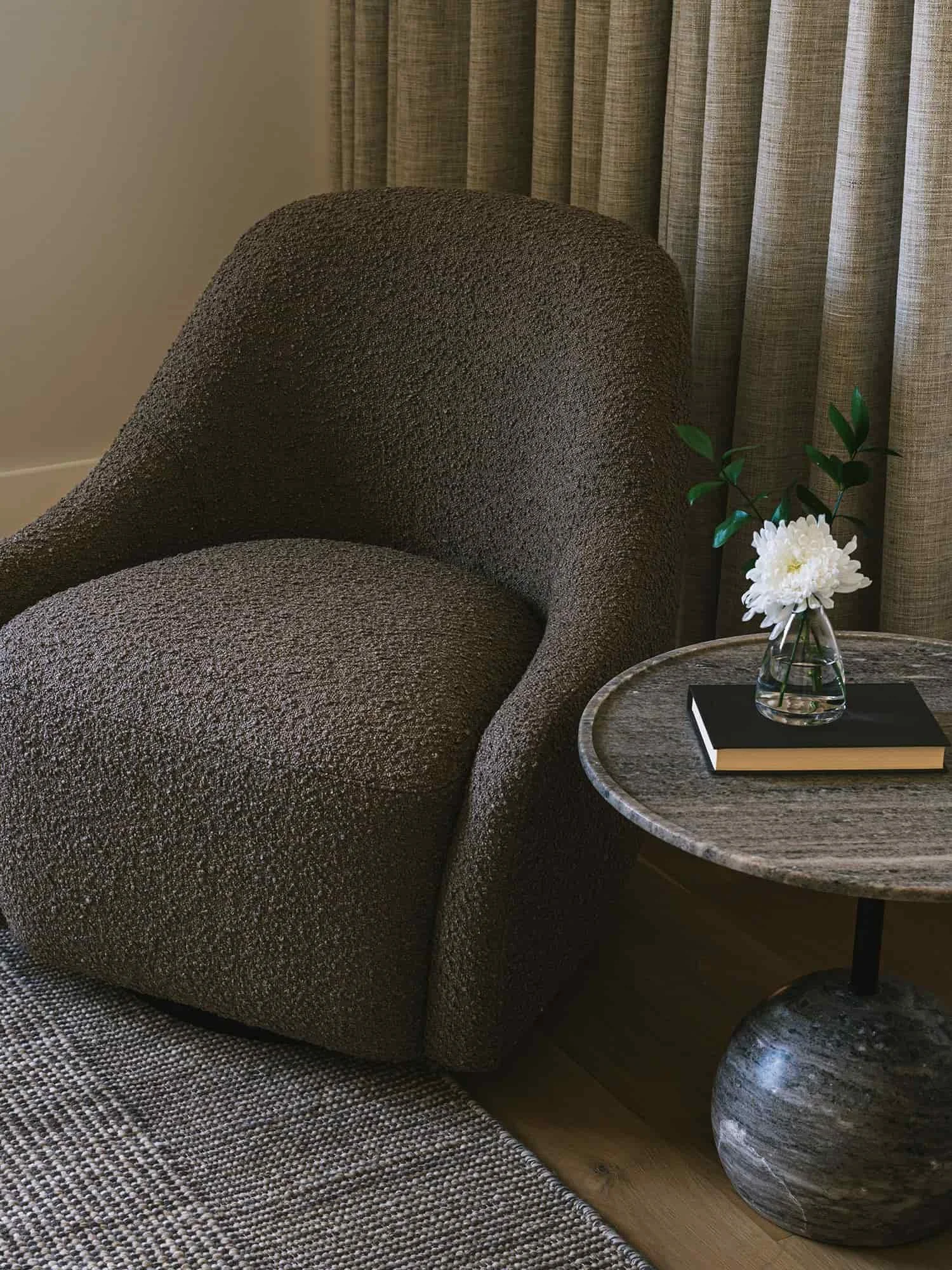 Sculptural upholstered lounge chair beside a round stone side table, with layered neutral textures and floor-to-ceiling drapery.