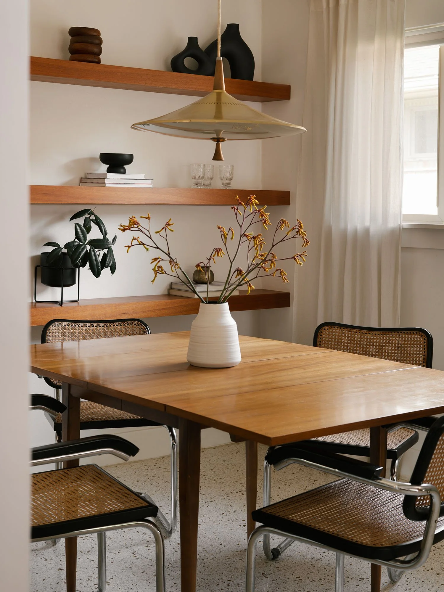 Authentic vintage Cesca chairs surrounding a wood dining table beneath a sculptural pendant, with styled floating shelves in the background.