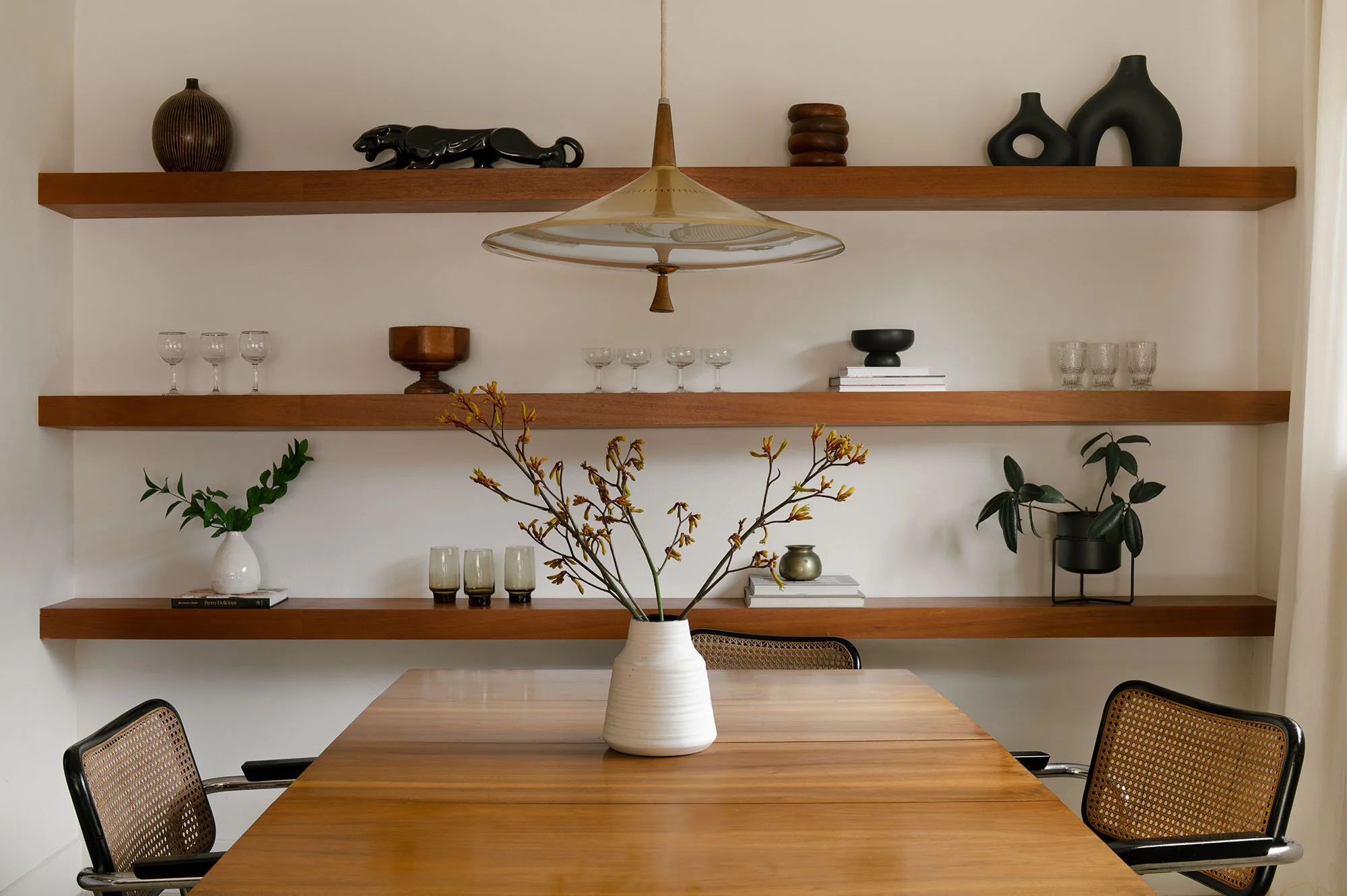 Mid-century modern dining room in Dallas featuring floating wood shelves, a sculptural vintage pendant light, cane-back dining chairs, and curated decorative accessories.