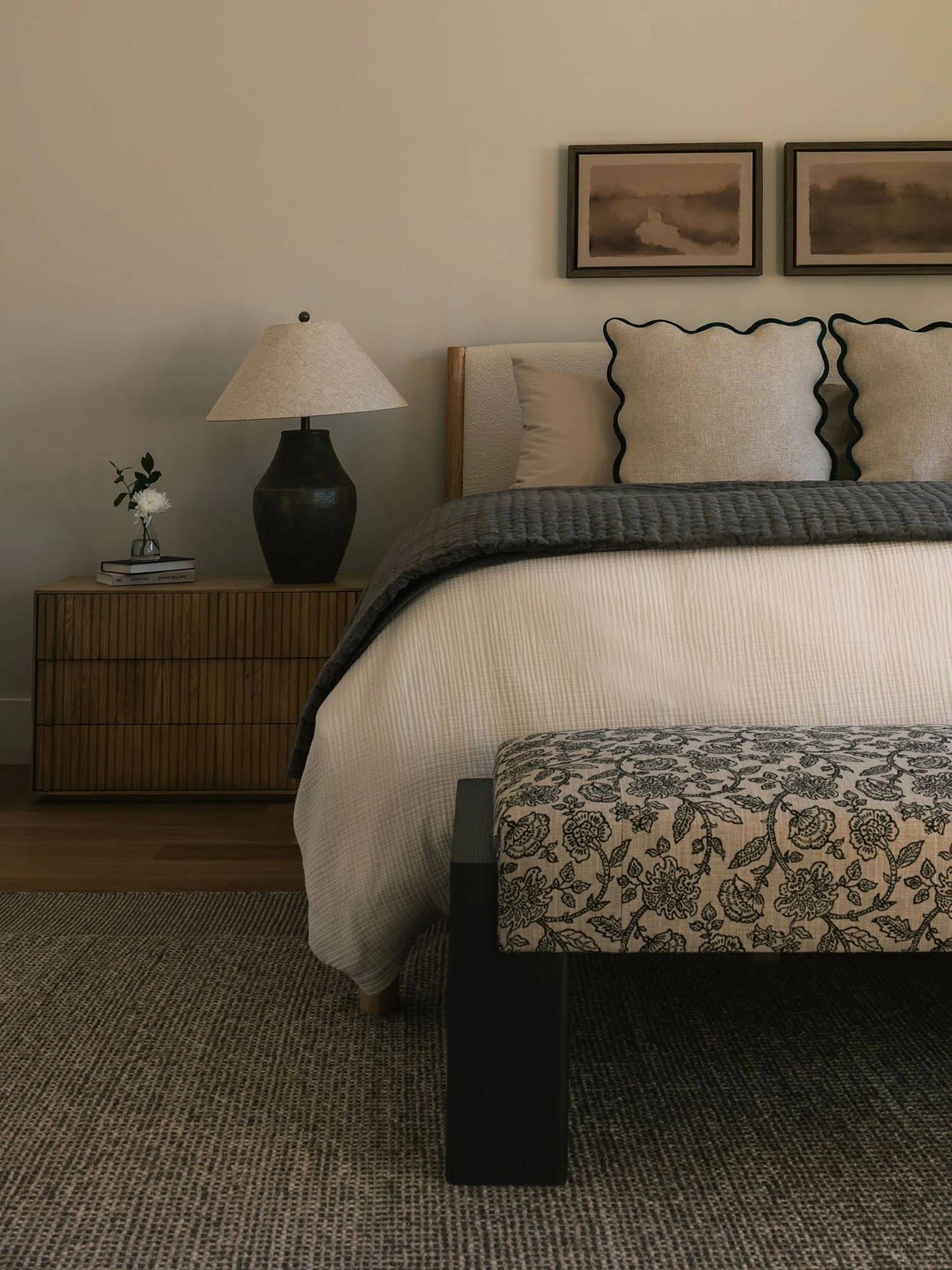Neutral bedroom featuring an upholstered bed, dark wood nightstand, sculptural table lamp, and floral-patterned bench.