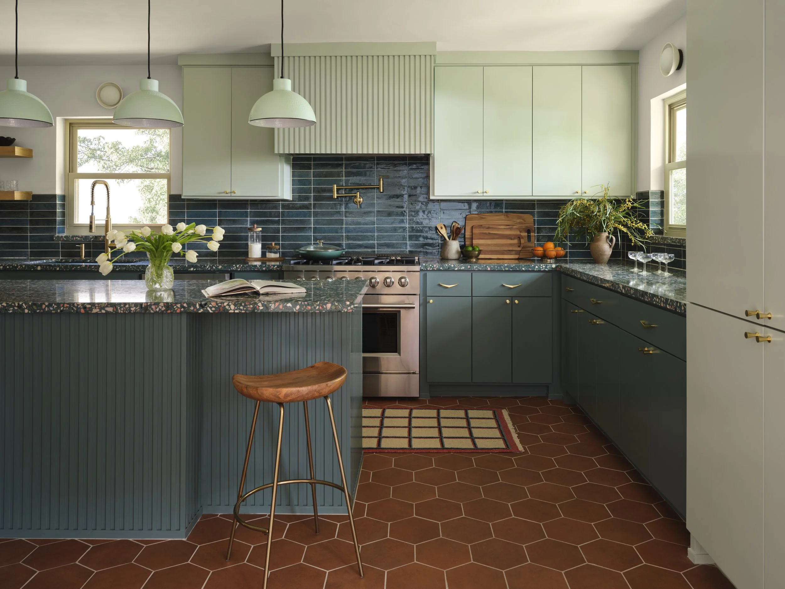 Colorful kitchen designed by Dallas interior designers at Paige Studio, featuring dark teal cabinetry, terrazzo countertops, blue tile, and terracotta hexagon flooring.