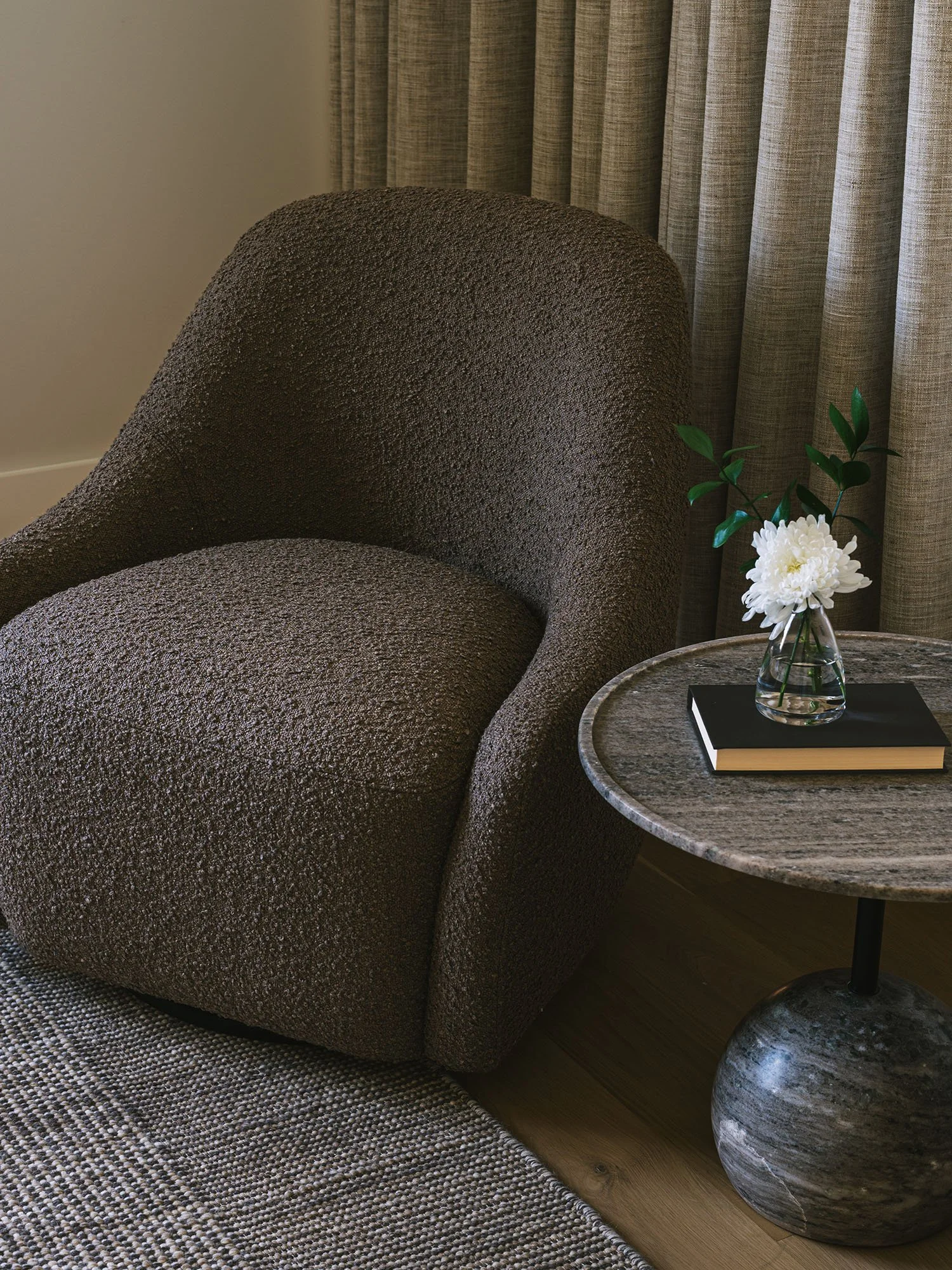 Dallas primary bedroom reading nook featuring a sculptural bouclé lounge chair, round stone side table, and textured floor-to-ceiling drapery.