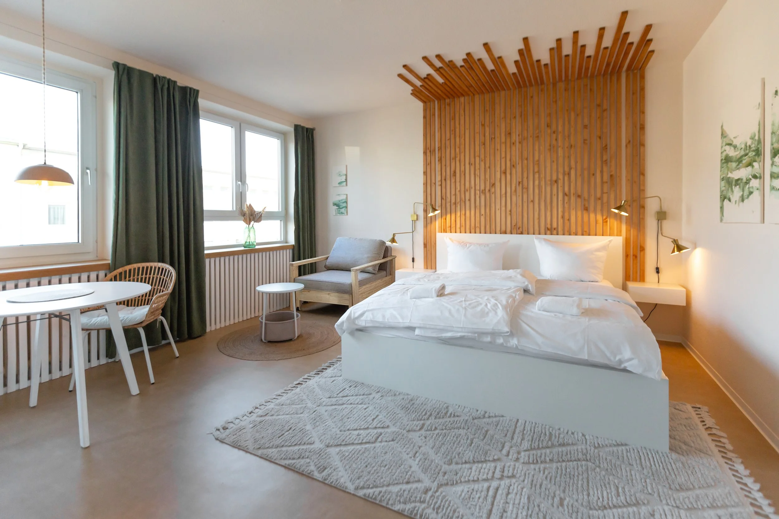 Schlafzimmer mit weihem Bett, Holz-Headboard, seitliche Nachttischlampen, Sitzecke mit Sofa, Esstisch und Stühlen, große Fenster mit grünen Vorhängen, Pflanze auf Fensterbank, Kunstwerke an der Wand.