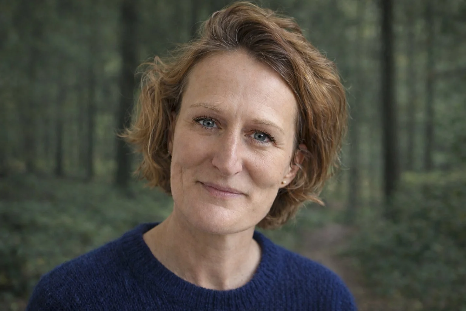 Close-up van een vrouw met korte, golvende, roodbruine haar in een bos met groene bomen.