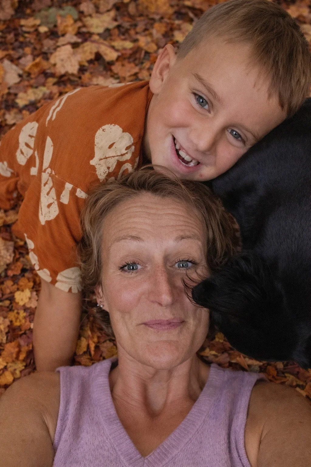 Een vrouw en een kind liggen op een bed van herfstbladeren, samen met een zwarte hond. De vrouw heeft rossig haar en draagt een lichtpaarse top. Het kind glimlacht breed en heeft blond haar, het ligt boven de vrouw en kijkt recht in de camera.