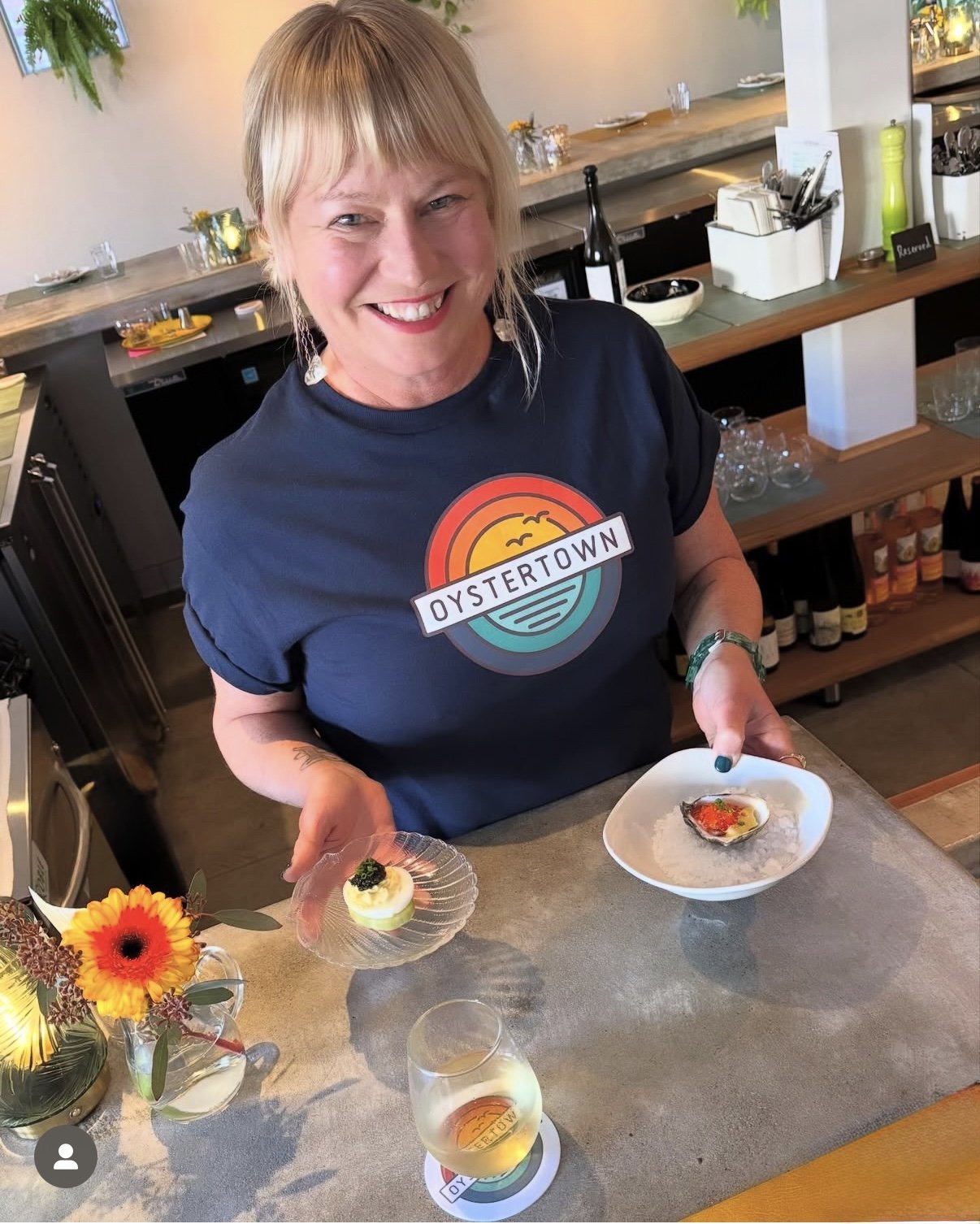 A woman smiling at the camera in a kitchen or bar area, holding a plate with two small gourmet dishes. She is wearing a navy blue T-shirt with a graphic that says 'Oystertown'. In front of her, there is a glass of wine and a small flower arrangement