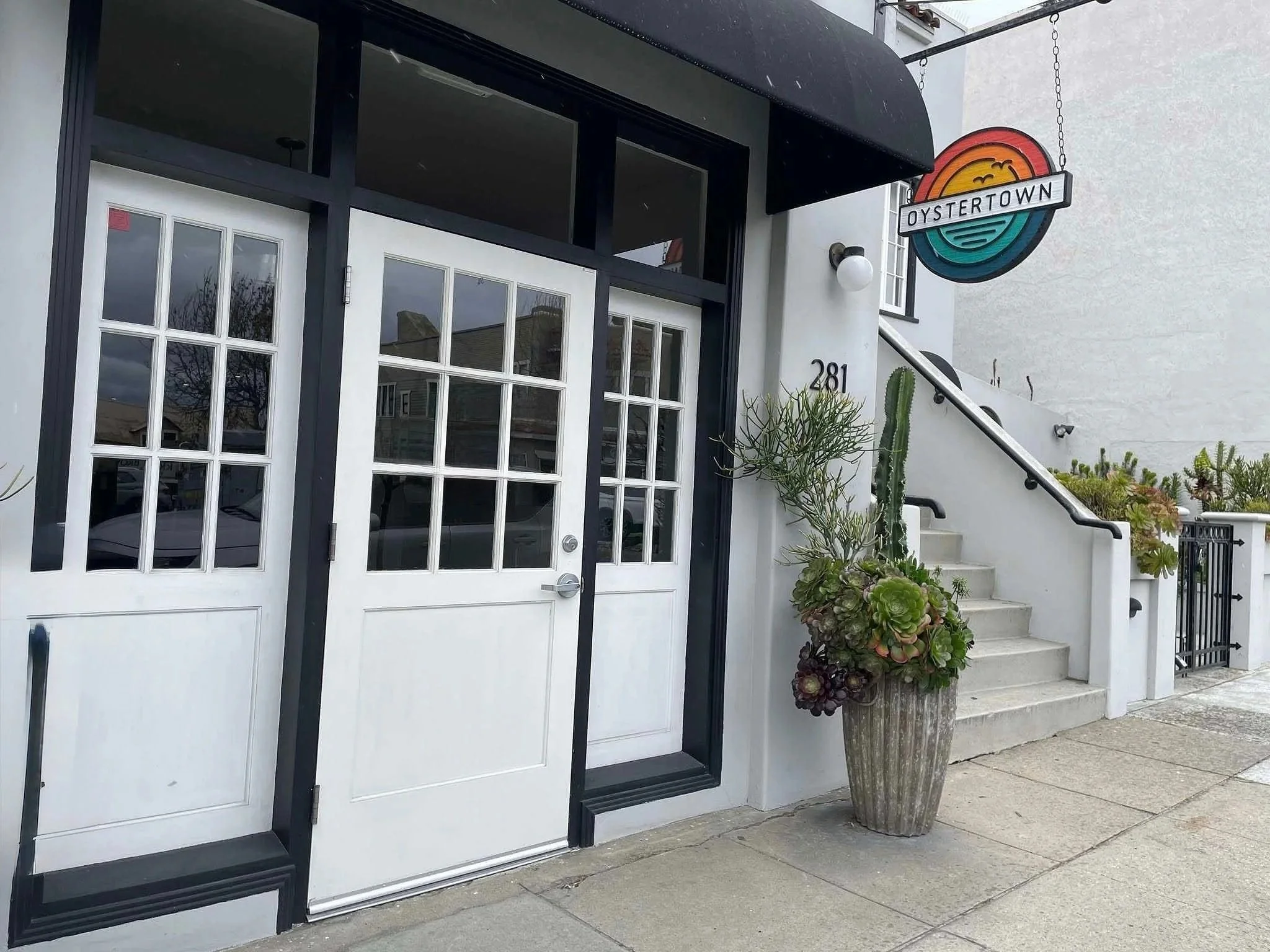Exterior view of a restaurant named Oystertown with white double doors, a potted cactus and succulents, a sign hanging above, and stairs leading to an upper level.
