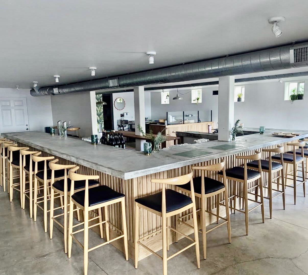 Modern, minimalistic bar with a large concrete countertop, surrounded by wooden barstools with black cushions, in a spacious, industrial-style interior with small high windows and exposed ductwork.