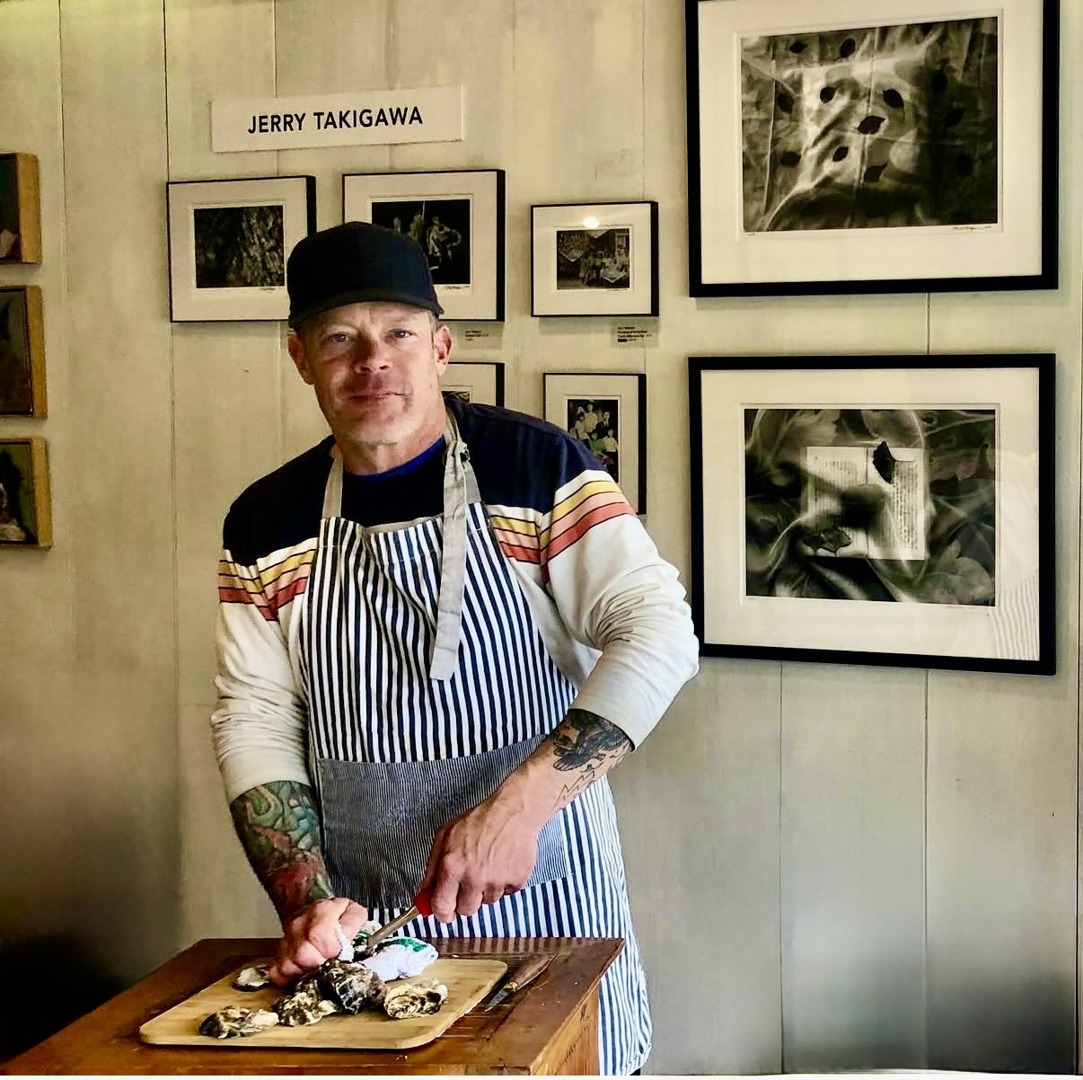 A man wearing a black hat, striped apron, and a long-sleeve shirt, standing at a wooden table with a cutting board and shellfish, in front of a wall decorated with framed photographs and artwork. The wall has a sign that reads 'JERRY TAKIGAWA'.
