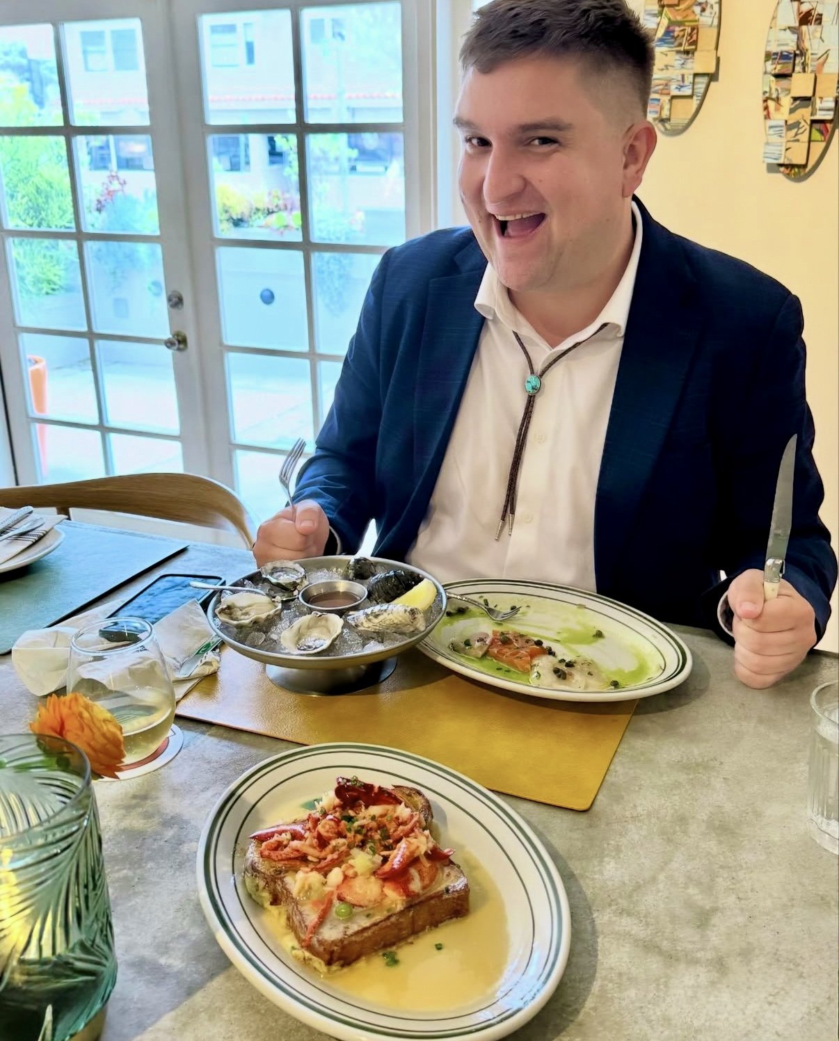 A man in a navy jacket and white shirt smiling at a table set with seafood dishes, including oysters, a lobster toast, and a plate with a green sauce, inside a restaurant near glass doors with a garden view.