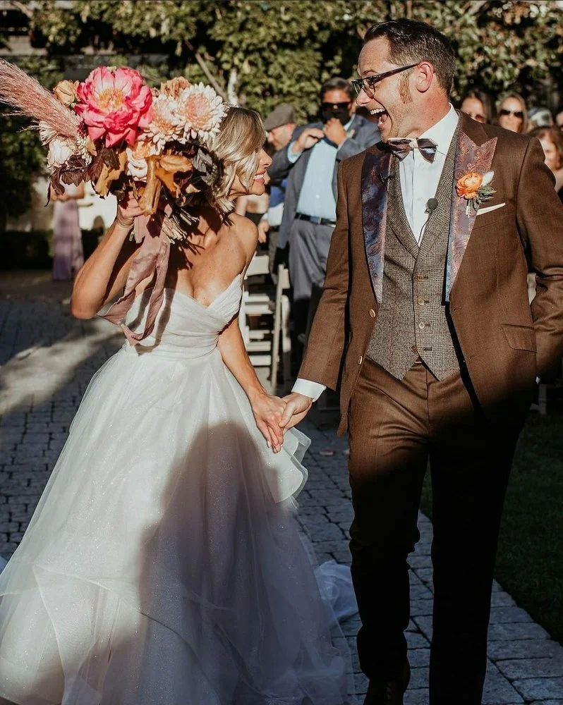 Bride in a white wedding gown holding a large bouquet of flowers, smiling at groom in a brown suit, outdoor wedding setting with guests in background.